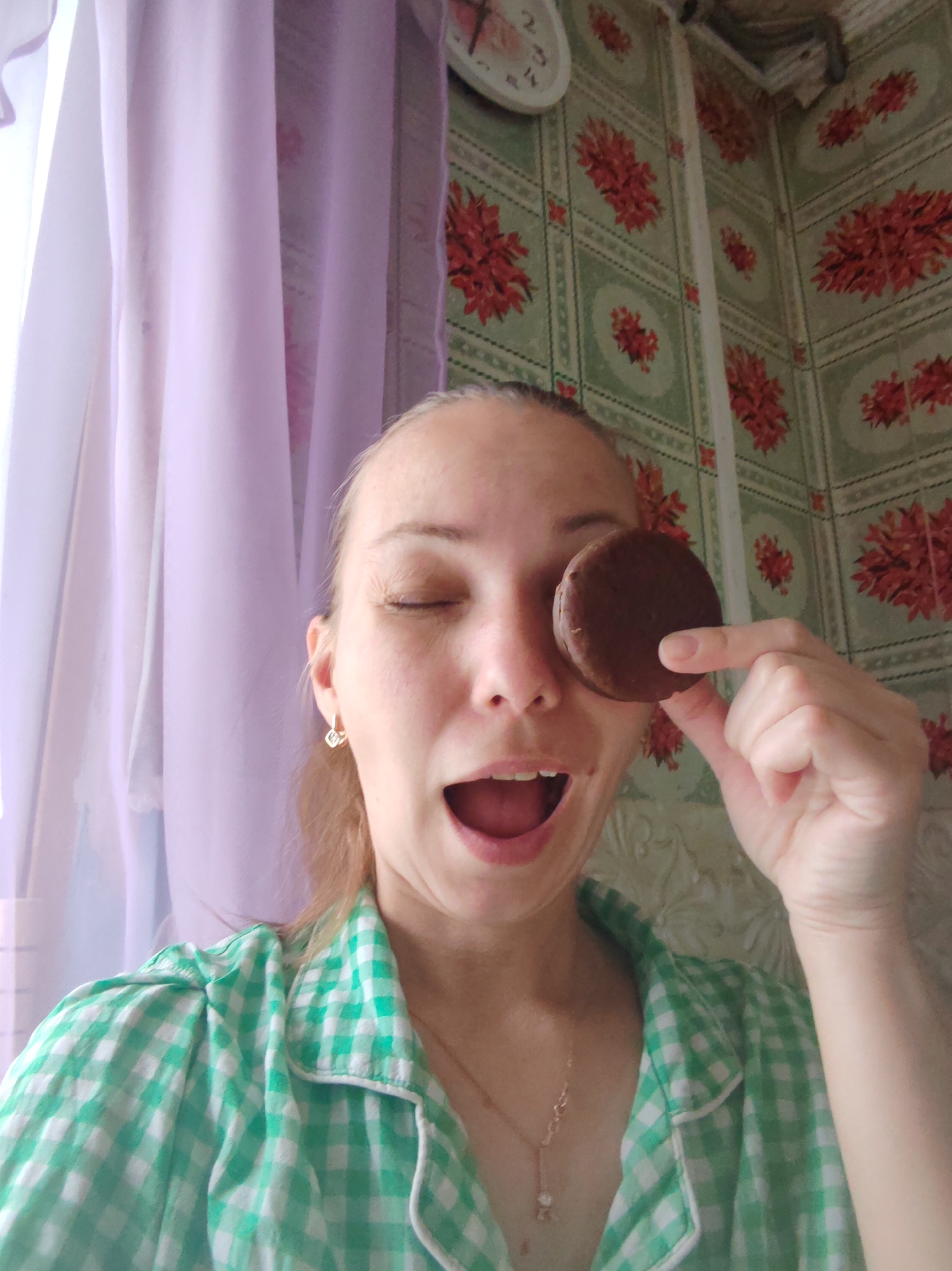 A woman with light skin and light brown hair is holding a chocolate pie up to her closed right eye, in front of a wall covered in green and red floral-patterned tile. She is wearing a green and white checkered shirt and has a gold necklace and earrings. Her mouth is open. A white clock is visible on the wall above and to the left of her head. To her left is a window with light purple curtains. The image was taken indoors, presumably in a kitchen. There are no discernible elements to confirm the location of the scene in the city of Чухлэм, Russia.