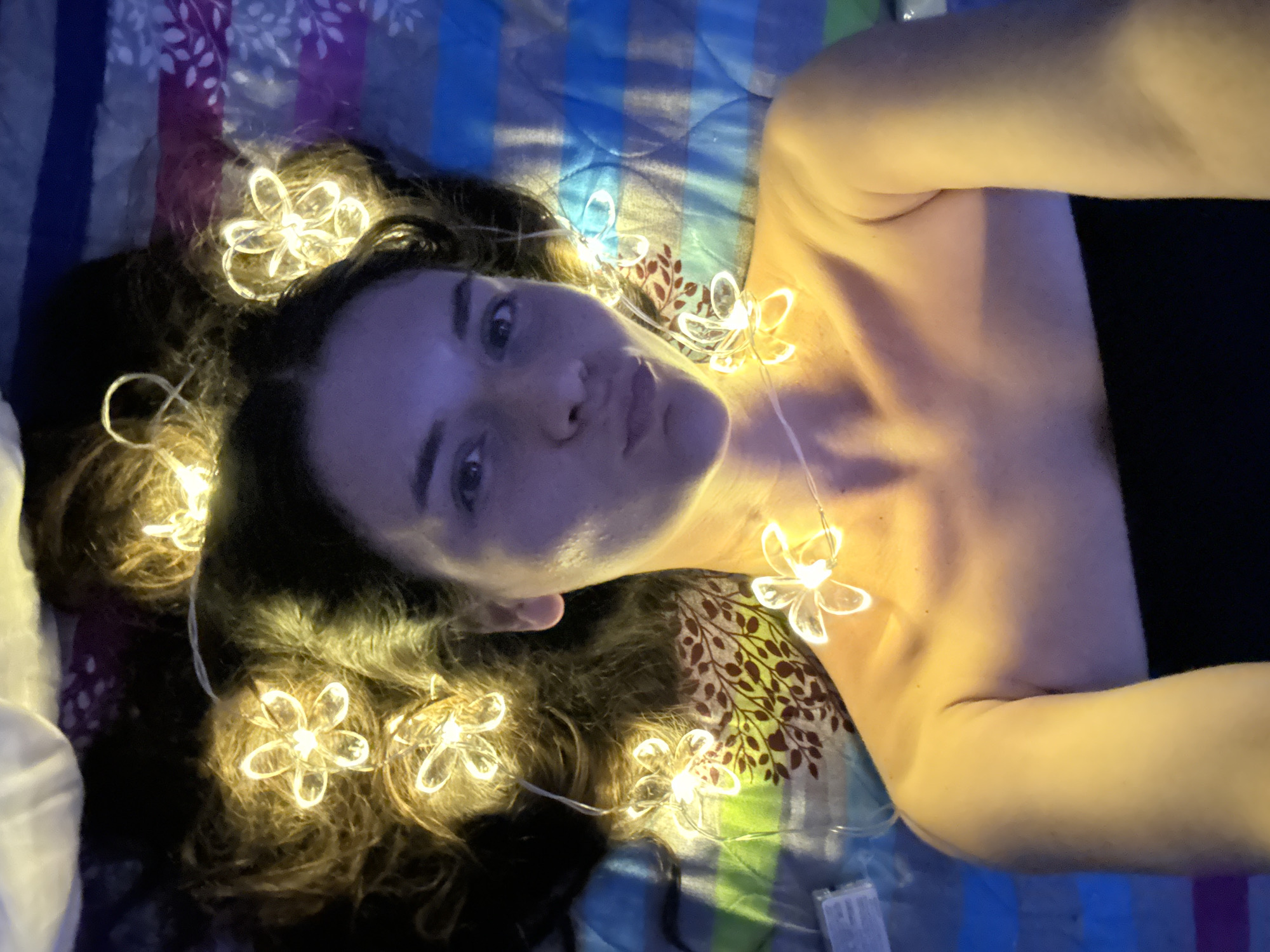 A woman with dark hair lies on her back on a bed with striped bedding. She is wearing a black strapless top. A string of flower-shaped lights is draped around her neck and into her hair, which is spread out around her head. She is looking at the camera with her lips puckered. The scene appears to be indoors. There is no discernible location within the city of Mũi Né, Vietnam.