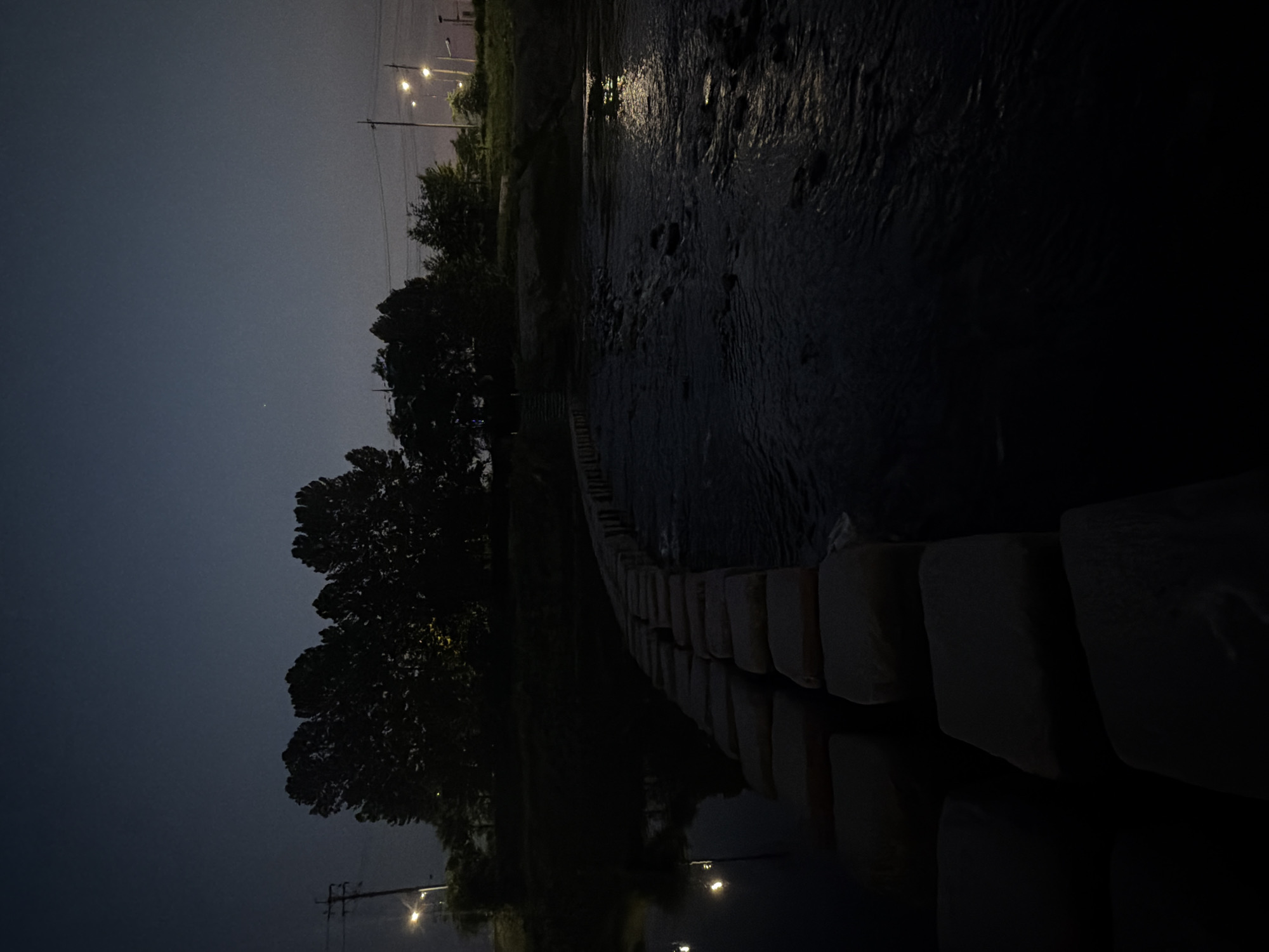 A low-angle, wide shot captures a serene night scene along a riverbank in Dongducheon-si, South Korea. The image is dominated by a dark, still river with scattered rocks. A row of large, square stepping stones forms a pathway across the water, leading the eye into the distance. The river's surface reflects the faint glow of distant streetlights and the dim twilight sky.
On the left bank, silhouetted trees stand tall against the dark sky, their shapes softened by the low light. A utility pole with visible streetlights is also present on the left side. On the right bank, more trees are visible, with more streetlights illuminating the area, suggesting a developed area or park. The overall atmosphere is tranquil and peaceful, with a sense of quiet solitude. There are no people or animals visible in the frame. The scene is likely captured during dusk or early night, given the presence of streetlights and the dim sky.