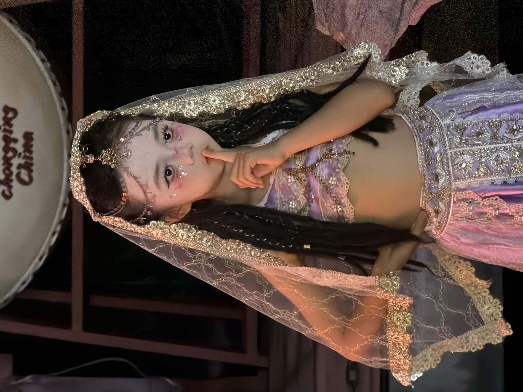 A young East Asian female, likely a child, is depicted in a costume with elaborate makeup and jewelry. She wears a sheer veil with gold trim, a sparkling purple and white top, and a long, layered purple skirt. Her hair is styled in braids, and she has glitter on her cheeks and around her eyes. She is holding a finger to her lips in a "shushing" gesture. The background appears to be a dark, indoor setting with some wooden structures visible. Text at the top of the image reads "chongqing China." This suggests the image was taken in Chongqing, China, which is located in Yuzhong District.