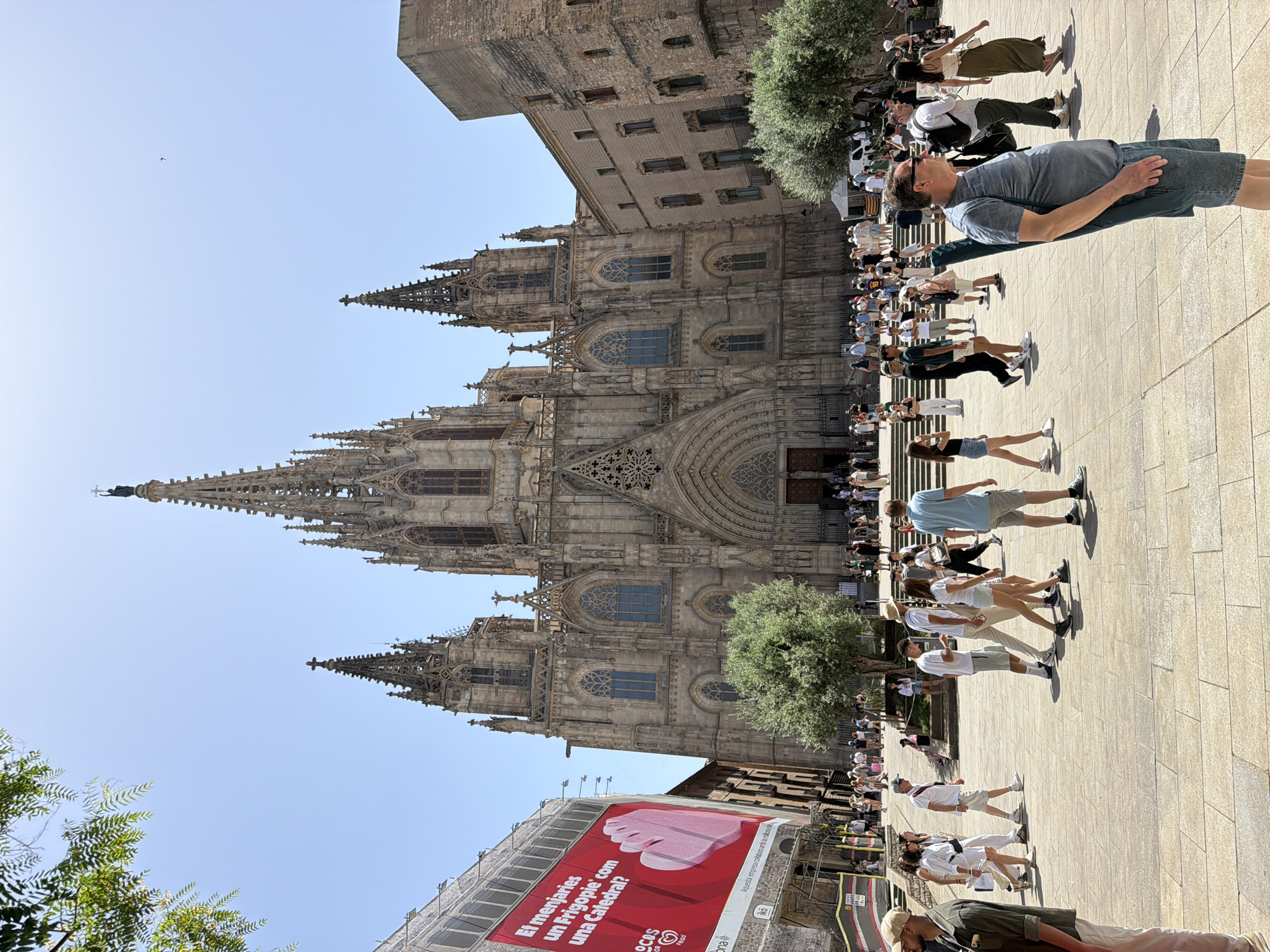 The image captures a sunny day in Barcelona, Spain, with the imposing Gothic architecture of the Barcelona Cathedral dominating the background. A group of people are gathered in the plaza in front of the cathedral, some appearing to be tourists taking in the sights, while others seem to be participating in a guided activity or demonstration. The people are dressed in casual summer attire, suggesting a warm and pleasant day. The sun casts long shadows on the stone pavement, indicating it might be late morning or early afternoon.
In the foreground, a large red banner is prominently displayed on a scaffolding, advertising a product called "Frigopie" with the question "El menjaries un Frigopie com una Catedral?" which translates to "Would you eat a Frigopie like a Cathedral?". This suggests a local marketing campaign or event. The overall atmosphere appears lively and communal, with people interacting and exploring the historic surroundings. The sky is a clear, bright blue, further emphasizing the fair weather conditions. The architectural details of the cathedral, with its intricate spires and ornate facade, are clearly visible, highlighting its grandeur.
