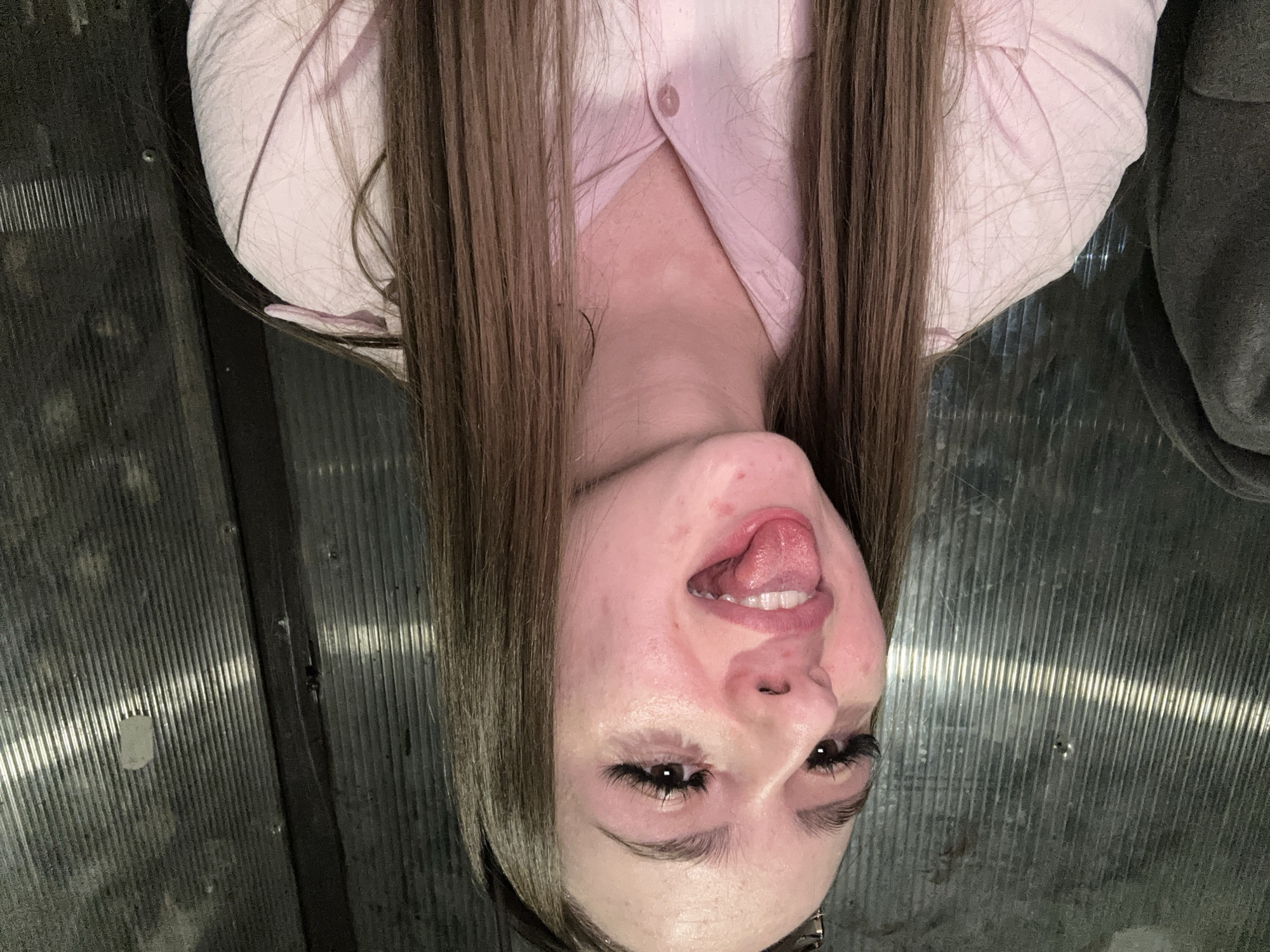 The image presents a headshot of a person, likely female, depicted in an inverted orientation. Her long, straight brown hair cascades from what would be the top of her head downwards in the frame. She is wearing a light pink collared shirt, partially visible, and a dark garment can be seen on the far right.
Her face is central, with her mouth open and tongue playfully sticking out. Her expression appears silly and lighthearted, with a direct gaze towards the viewer. She has prominent eyelashes, possibly extensions, and some skin redness is visible on her cheeks and forehead.
The background features a textured, grey, ribbed or corrugated metallic surface. A dark, vertical seam or structural element runs through this background. The lighting is dim, suggesting an indoor setting or low-light conditions. There is no visible text.