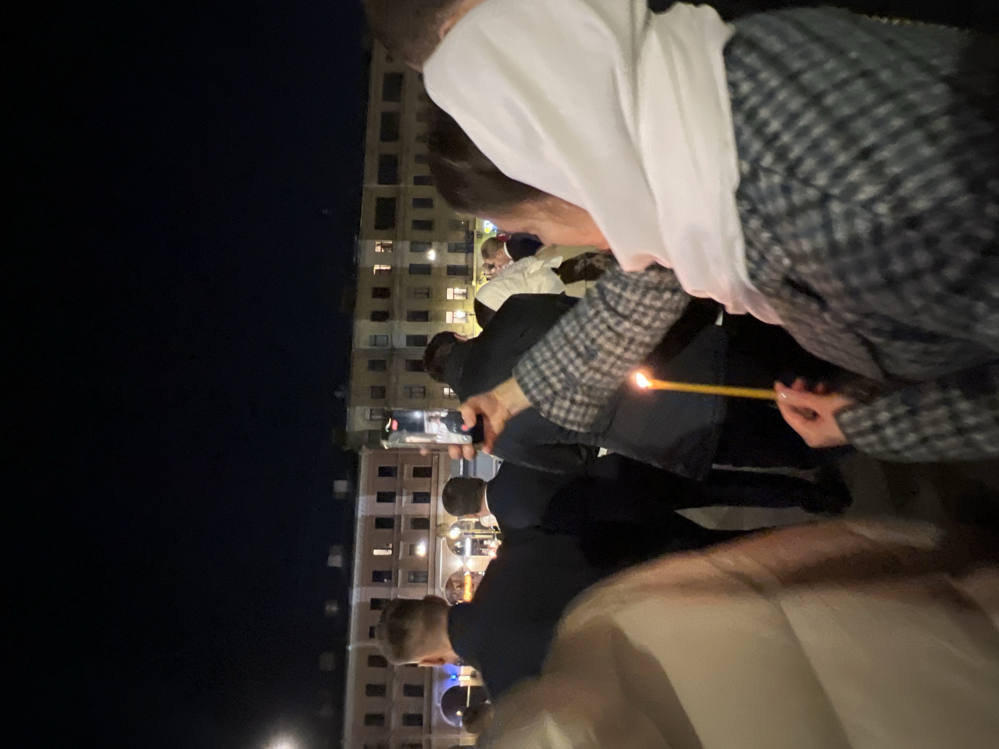 A nighttime scene in Saint Petersburg, Russia, features a group of people, likely gathered for a religious event. Several individuals are holding lit candles. One person is using a smartphone to record the scene. A tall building with illuminated windows serves as the backdrop. The scene is viewed from a close-up perspective, with individuals appearing in the foreground, one wearing a white head covering and a plaid jacket.