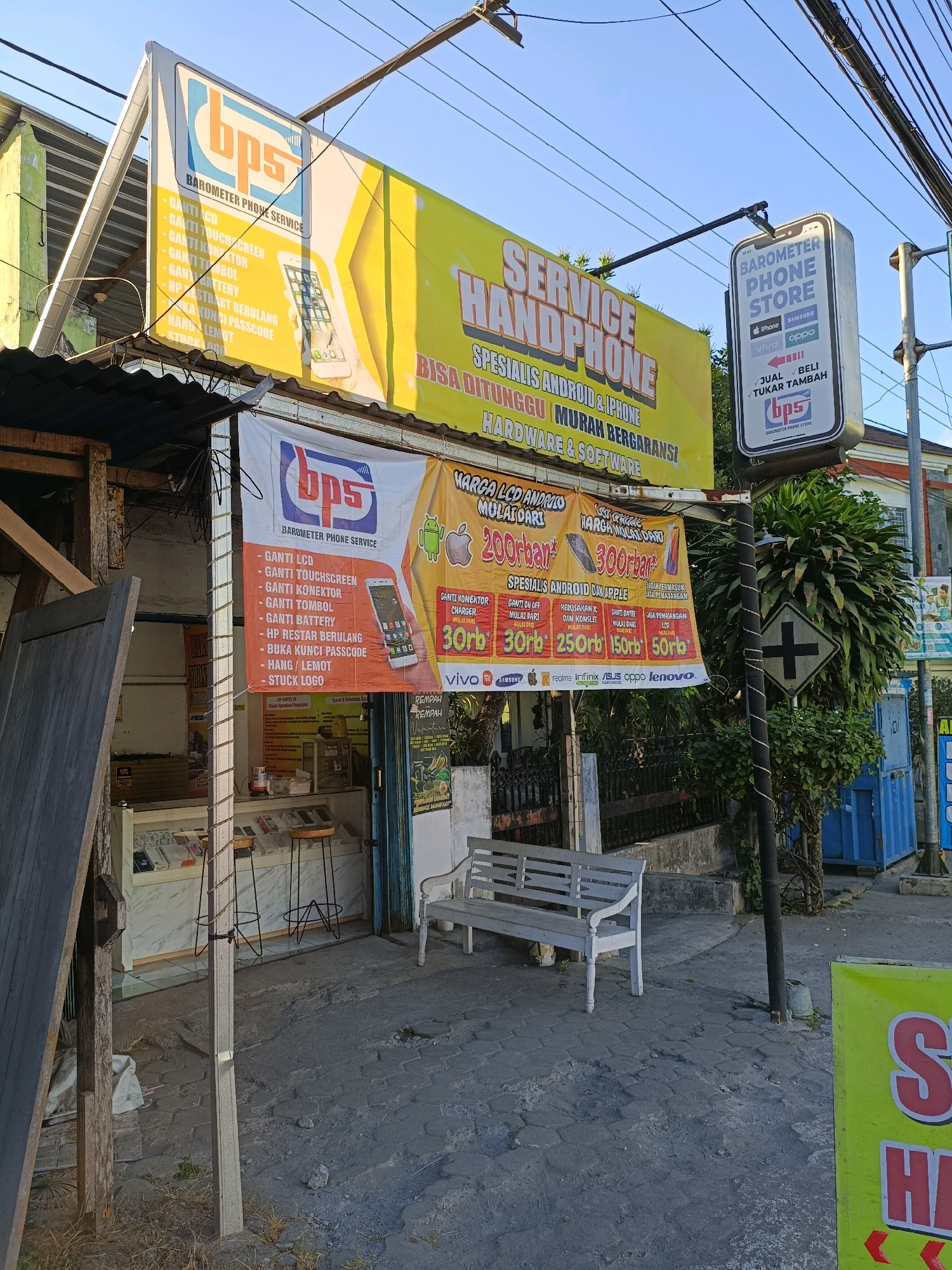 The image depicts a storefront in Maguwoharjo, Indonesia, specializing in phone services and sales. The facade features several prominent signs. A large yellow banner reads "SERVICE HANDPHONE SPESIALIS ANDROID & IPHONE BISA DITUNGGU | MURAH BERGARANSI HARDWARE & SOFTWARE". Above this, a white sign with the "bps" logo advertises "BAROMETER PHONE SERVICE" and lists services like "GANTI LCD", "GANTI TOUCHSCREEN", "GANTI KONEKTOR", "GANTI TOMBOL", "GANTI BATTERY", "HP RESTART BERULANG", "BUKA KUNCI PASSCODE", "HANG / LEMOT", and "STUCK LOGO". A vertical sign shaped like a smartphone, also featuring the "bps" logo, states "BAROMETER PHONE STORE" and lists brands like iPhone, Samsung, Vivo, and Oppo.
Below the overhead signs, a white banner displays the "bps" logo and lists similar phone services. Additional smaller banners advertise "HARGA MULAI DARI" (prices starting from) 200rb for LCD and 300rb for other services, along with specific prices for services like "GANTI KONEKTOR", "CHARGER", "GANTI BATERAI", "HARDWARE & SOFTWARE", and "HP PEMASANGAN". Various phone brands are listed, including Vivo, Samsung, Realme, Infinix, Asus, Oppo, and Lenovo.
The shop interior is visible through a glass display counter showcasing mobile phones. Two high stools are positioned in front of the counter. Outside the shop, a white wooden bench sits on a paved pathway. The environment appears to be a street with power lines overhead and some greenery, including a large bush on the right. The overall scene suggests a commercial area on a clear day.