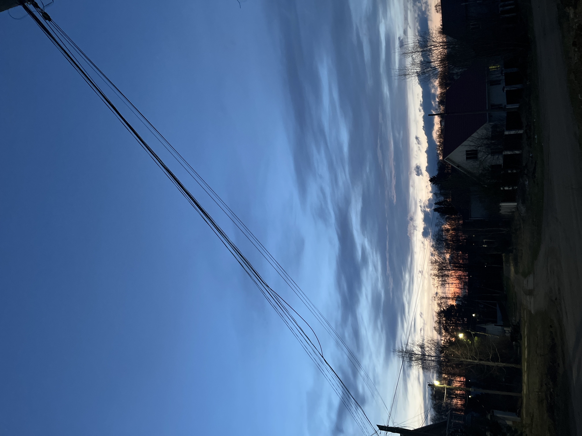 The image captures a tranquil rural or suburban scene in Chaikovsky, Russia, during either dusk or dawn. The sky dominates the upper portion of the frame, transitioning from a deep, clear blue in the upper left to lighter blues and streaky grey clouds towards the horizon on the right. A distinct band of orange and pink light along the distant horizon indicates the setting or rising sun, creating a vibrant contrast with the cooler sky.
Multiple black power lines stretch diagonally across the upper-left quadrant of the sky, attaching to a utility pole partially visible at the far left edge. The lower half of the image features a landscape of dark silhouettes. A dense line of mostly bare trees forms an irregular horizon line. Below the trees, several houses are visible as dark shapes with discernible rooflines and a few illuminated windows, suggesting a residential area. At least two streetlights cast a warm, yellowish glow, providing artificial illumination against the fading natural light. A dark strip of ground, possibly a road or path, is visible at the very bottom right.
No people or specific activities are depicted, contributing to the serene and quiet atmosphere of the moment. There is no visible text in the image.