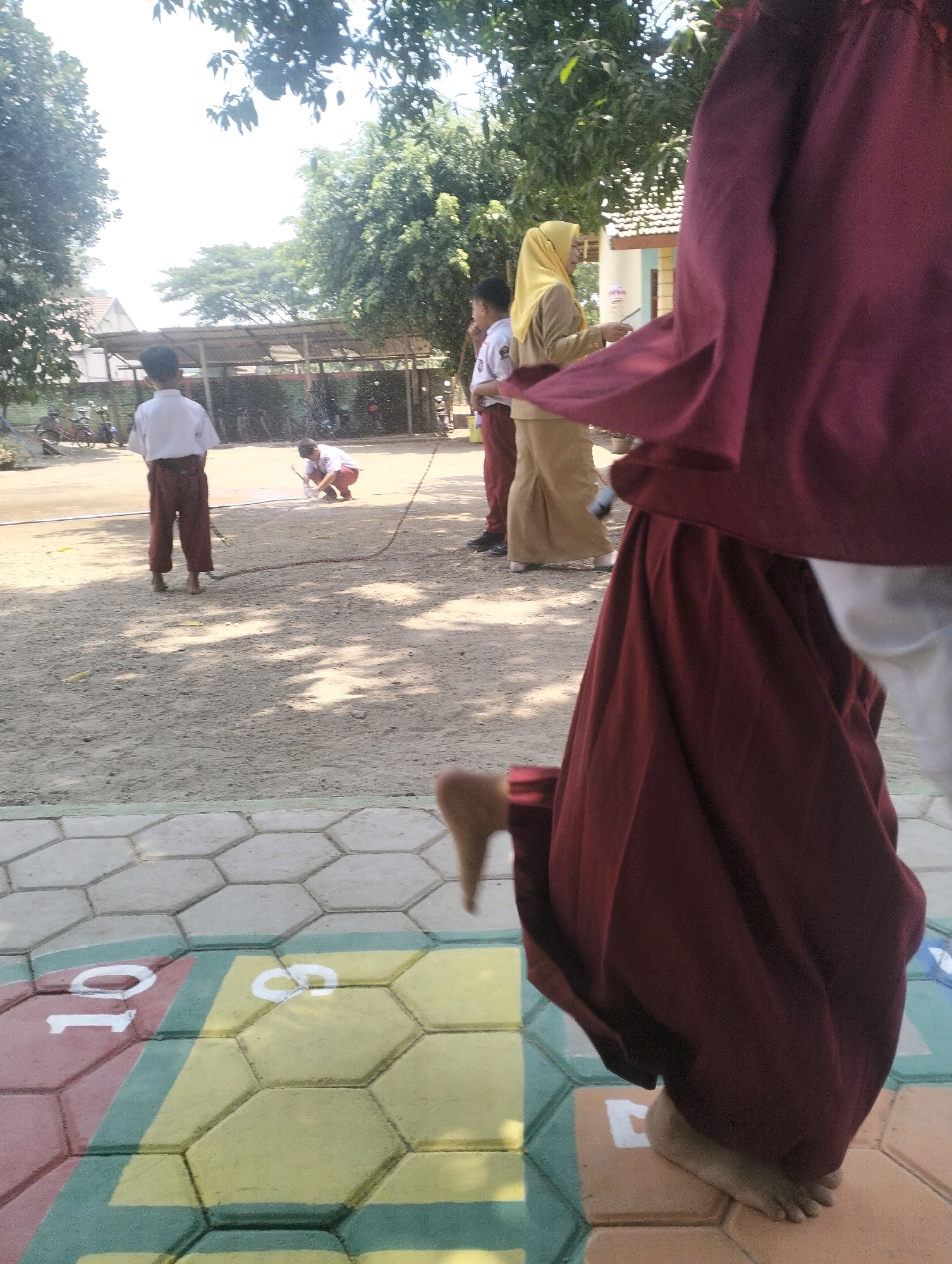 In the city of Blabakan, Indonesia, children are engaged in an outdoor activity. In the foreground, a child's foot is visible stepping on a colorful hopscotch grid. The grid consists of hexagonal tiles painted in red, yellow, and green, with white numbers indicating squares for jumping. Behind this, a group of children are gathered. One child, wearing a white shirt and brown pants, stands facing away from the camera. To their left, another child is crouched down, holding a stick, seemingly participating in a game or task. Further back, an adult wearing a yellow hijab and light brown attire is seen speaking to another child in a white shirt and brown pants. The background reveals trees, a building with a tiled roof, and a shaded area with what appear to be motorcycles parked. The ground is covered in sand and dirt, with patches of shade cast by the surrounding trees. The scene suggests a schoolyard or a community play area.