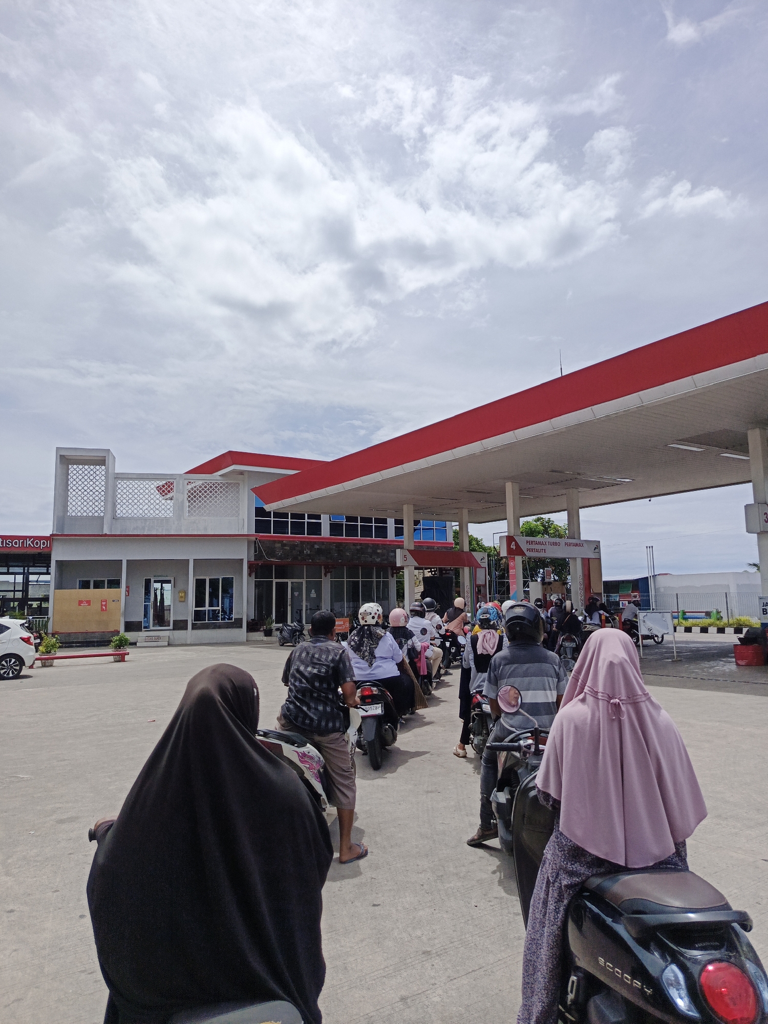 This is a wide-angle, eye-level shot of a queue of motorcyclists at a gas station in Cotbadatunong, Indonesia. The gas station has a red and white canopy supported by white pillars. In the background, there is a building with white and grey elements, and some signs. The sky is overcast with white clouds. The foreground features a line of motorcycles with riders, most wearing helmets. Two individuals in the immediate foreground are riding motorcycles and are partially visible from behind, one wearing a black hijab and the other a pink hijab. The motorcyclists are lined up waiting to refuel. The ground is paved concrete.