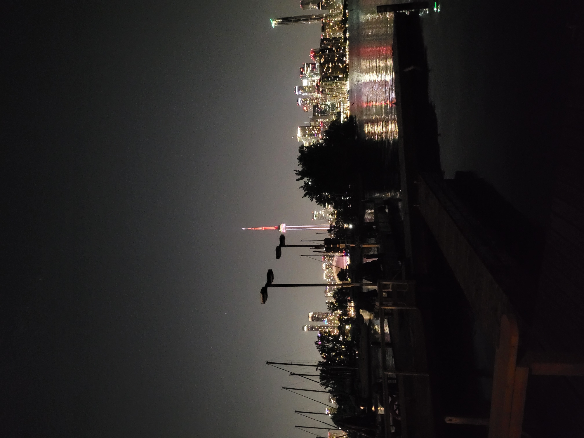 This is a nighttime photograph of the Toronto skyline as viewed from the water. The prominent CN Tower, illuminated with red and white lights, stands out against the dark sky. The city skyline is dotted with numerous illuminated buildings, reflecting on the calm water in the foreground. On the left side of the image, several sailboats are docked, their masts silhouetted against the city lights. In the foreground, a wooden dock or pier is visible, with some benches and other objects casting dark shadows. The overall atmosphere is serene and urban.