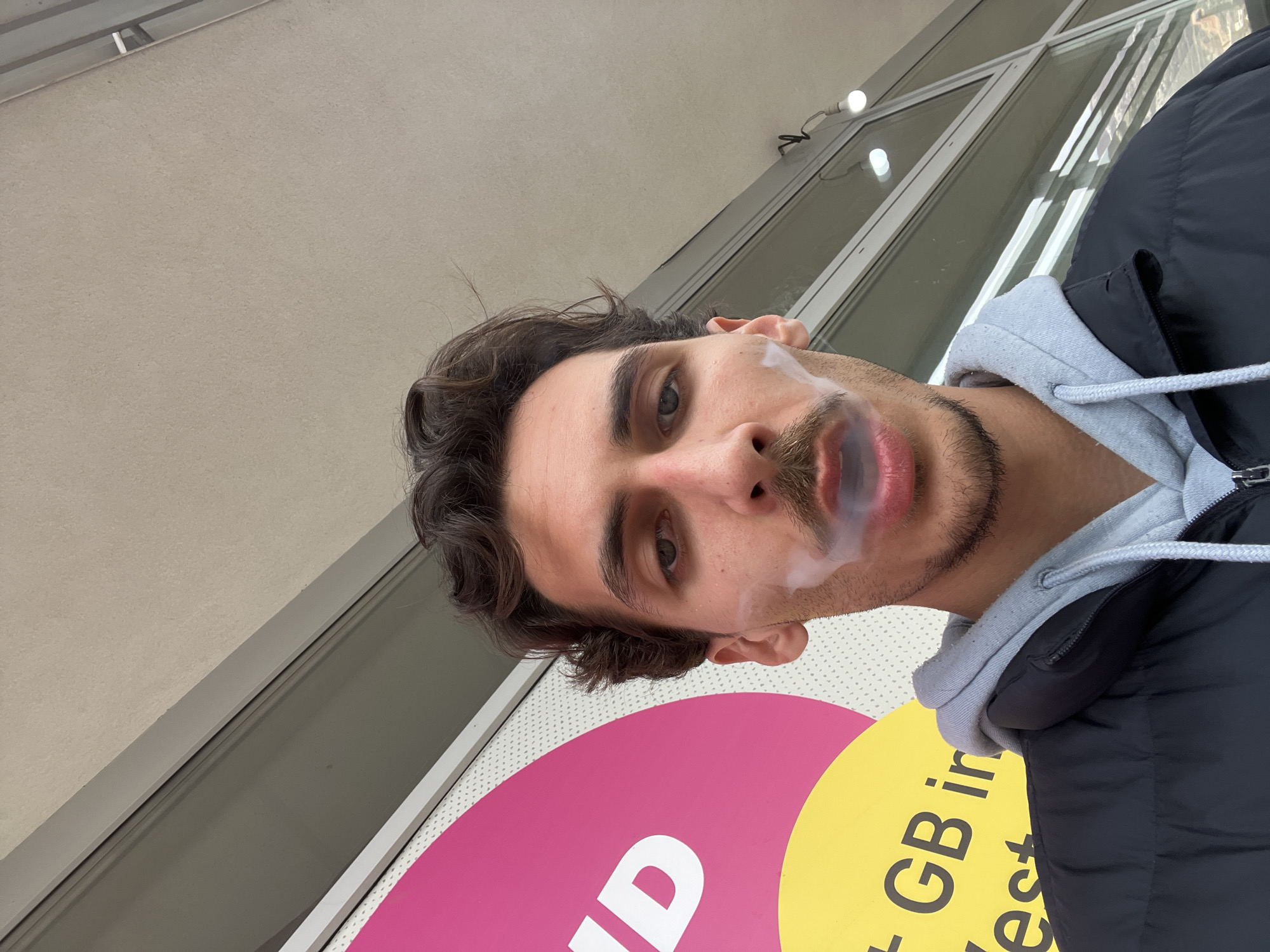 A selfie of a man with brown curly hair, light eyes, and a trimmed beard exhaling smoke, taken from a low angle. He is wearing a grey hooded sweatshirt beneath a black puffy jacket. The background includes a beige building facade, a multi-pane window, and a sign with pink, yellow, and white colors. The sign includes letters that are partially visible; they appear to be part of the word "QUID" and the letters "GB" are also visible on the sign. The setting appears to be urban, potentially within the city of Budva, Montenegro.
