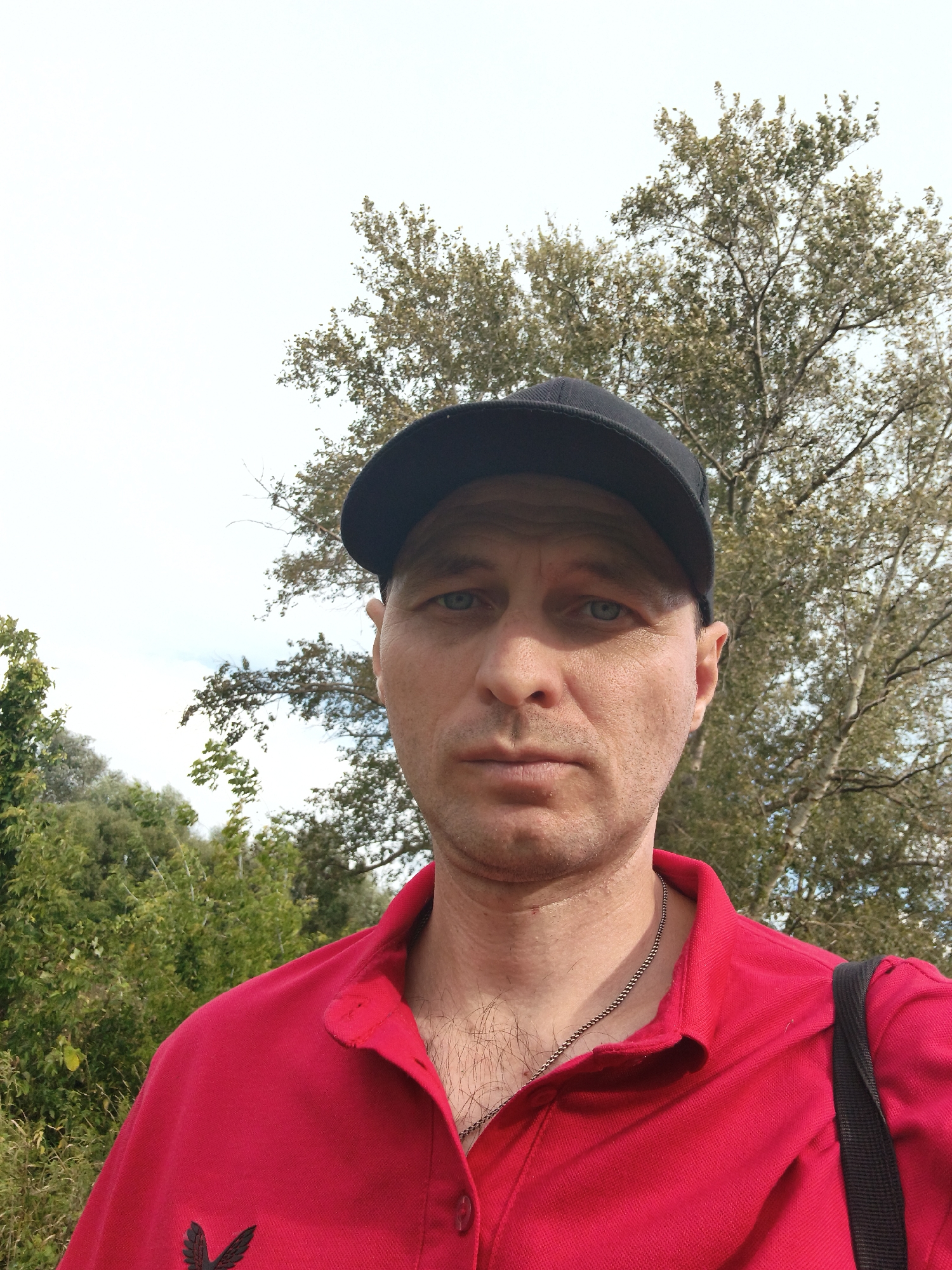 A man wearing a red polo shirt and a black baseball cap is photographed outdoors in a natural setting. He has light blue eyes and a neutral expression. A silver chain is visible around his neck. Behind him, there are green trees and a pale sky. The man is positioned slightly off-center to the right of the frame. There are no discernible city landmarks or structures visible. The image appears to be a selfie taken in a rural or park-like environment, possibly outside of Mokshan, Russia, though no specific location details are evident from the visual information.