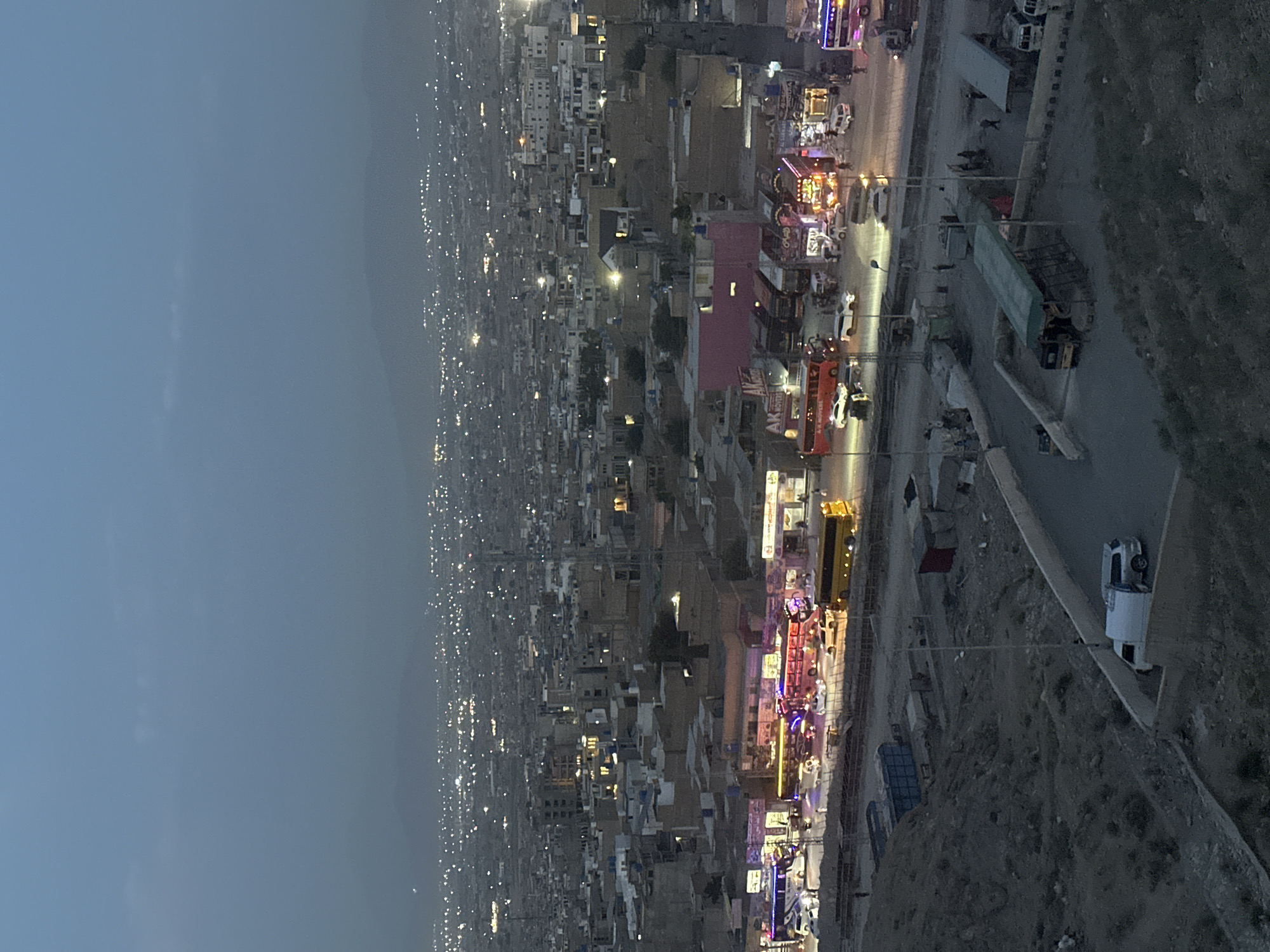 The image shows a nighttime view of a portion of Quetta City Tehsil, Pakistan, observed from an elevated perspective. A densely populated urban area stretches into the distance with numerous buildings and lit windows, suggesting a high concentration of residents. In the foreground and extending toward the middle ground is a street lined with shops and businesses, characterized by brightly lit storefronts. Vehicles, including buses and cars, are visible along the street, indicating ongoing vehicular activity. On the right side of the image, a hillside or embankment borders the road. The sky appears to be overcast or hazy, with a muted color gradient.