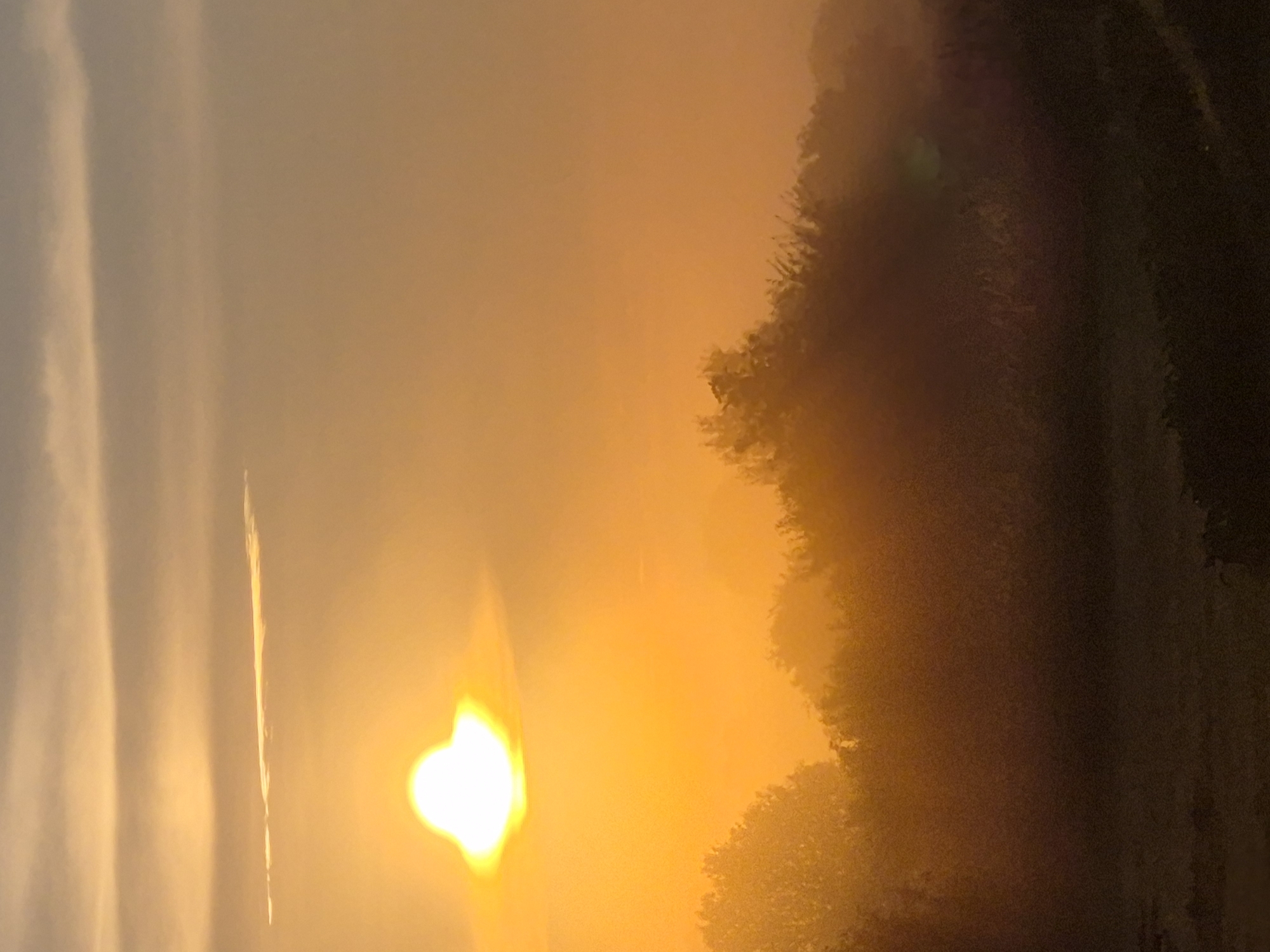 The image depicts a landscape scene at dawn or dusk. The sun, a bright, luminous orb with a halo effect, is positioned in the upper left quadrant of the frame. Its light casts a warm, orange glow across the sky and illuminates a thin, horizontal cloud strip above it. The lower half of the image is dominated by a silhouetted treeline, shrouded in a soft haze or mist. The foreground appears to be a darker, less distinct area, possibly fields or ground cover. The scene evokes a serene and slightly ethereal atmosphere, characteristic of sunrise or sunset in a rural or natural setting. The location in Sooblase, Estonia, is not visually identifiable from the image itself.