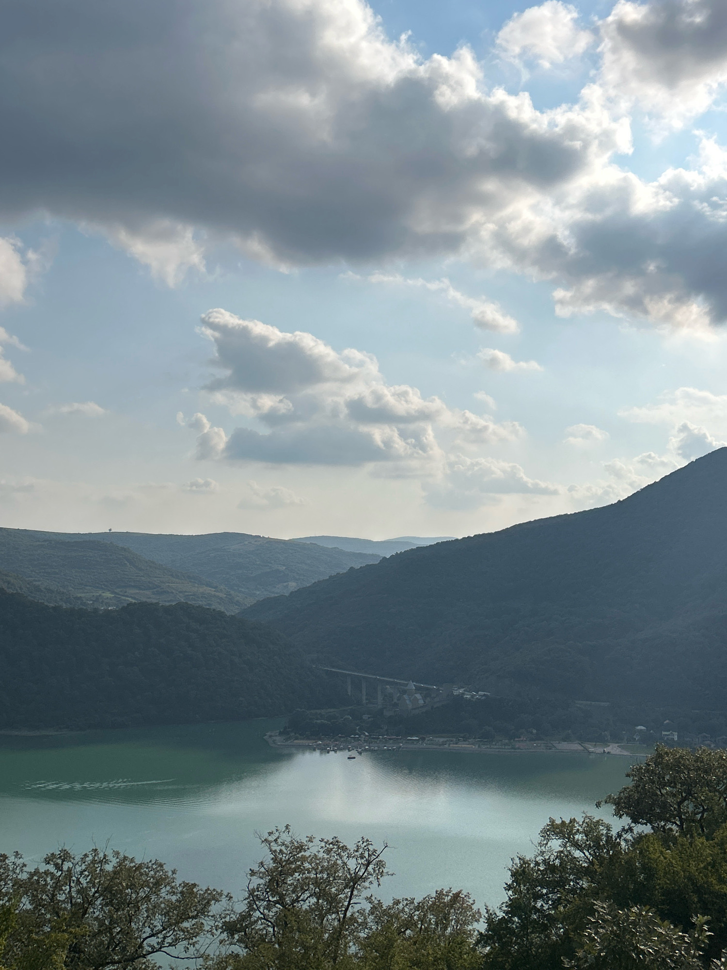 This is a vertical, eye-level, full shot of a wide, greenish lake reflecting the sky and surrounded by rolling, tree-covered hills. The sky is filled with dramatic, gray and white clouds, with patches of bright blue visible. In the distance, a bridge spans across the lake, and there appear to be buildings and boats along the shoreline. The foreground is dominated by the dark silhouettes of leafy trees. The scene appears to be a natural landscape with some man-made structures. The location is identified as being in Shalikiantkari, Georgia.