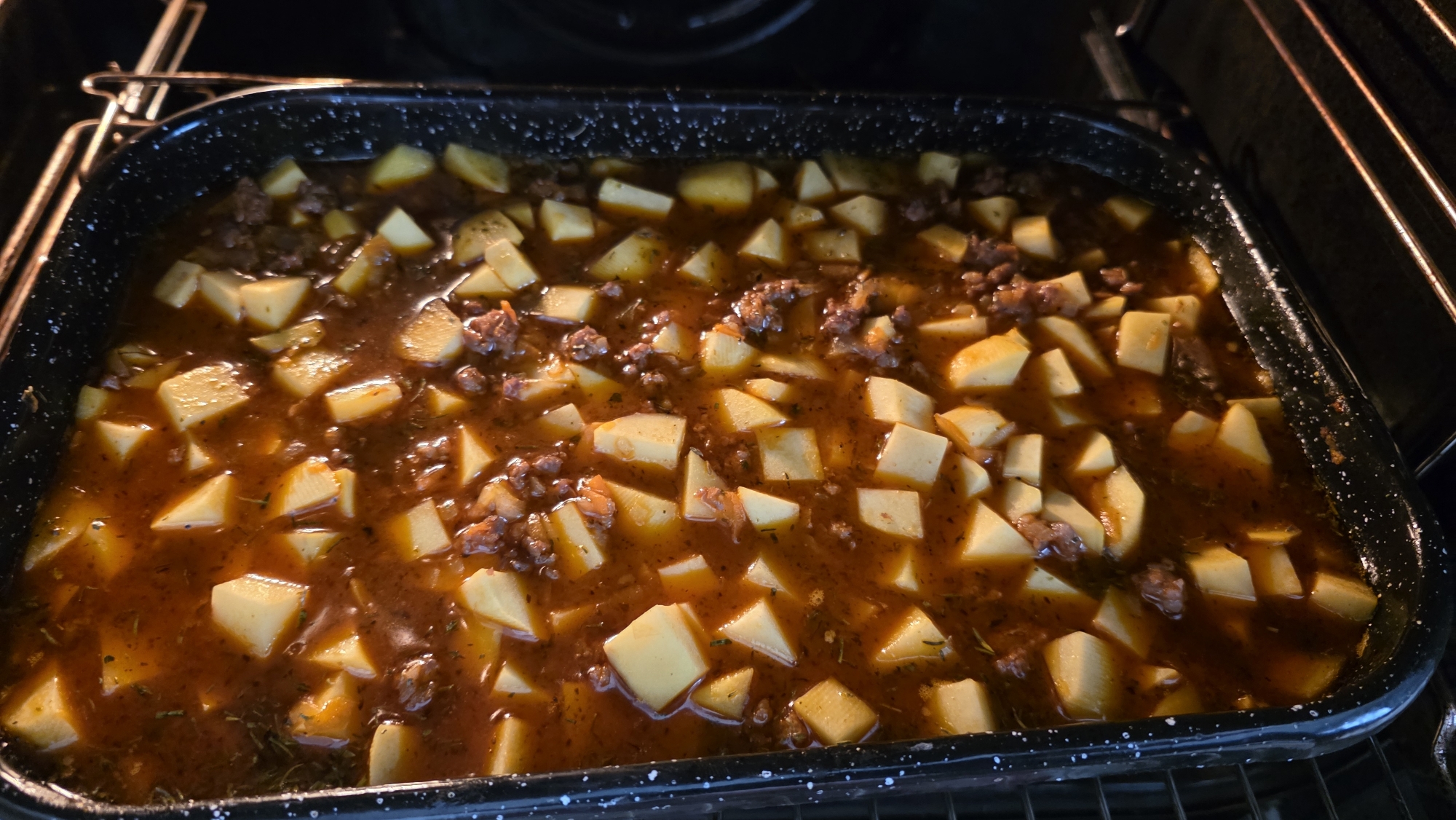 A black baking pan filled with a stew-like dish sits inside an oven. The dish appears to contain diced potatoes, ground meat, and a brownish sauce. The oven racks are visible. The context of Blagoevgrad, Bulgaria, is not evident in the image.