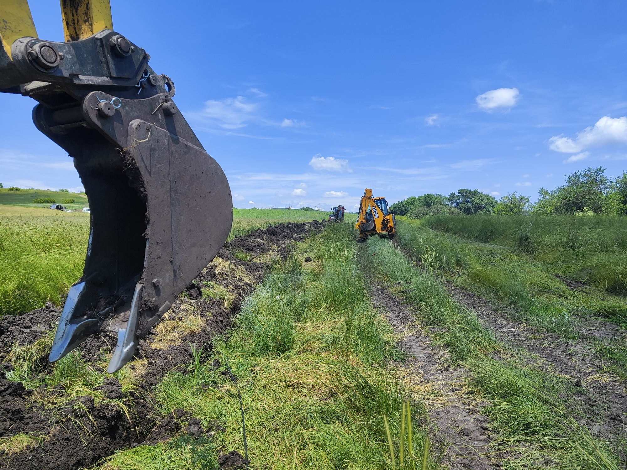 The image depicts an outdoor scene in Malak Manastir, Bulgaria, on a clear day with a blue sky and scattered white clouds. In the foreground, the dark, muddy bucket of an excavator is prominently positioned, angled downwards as if in the process of digging. The excavator arm is yellow and black. Behind the excavator, a trench is being dug through a grassy field. Another yellow excavator, with the brand "JCB" visible, is working further down the trench. The field is covered in tall green grass, with some areas appearing matted and disturbed by machinery. In the distance, there are trees lining the horizon and a few small white structures are visible in the far background on the left. The overall impression is of ongoing construction or land alteration work in a rural setting.