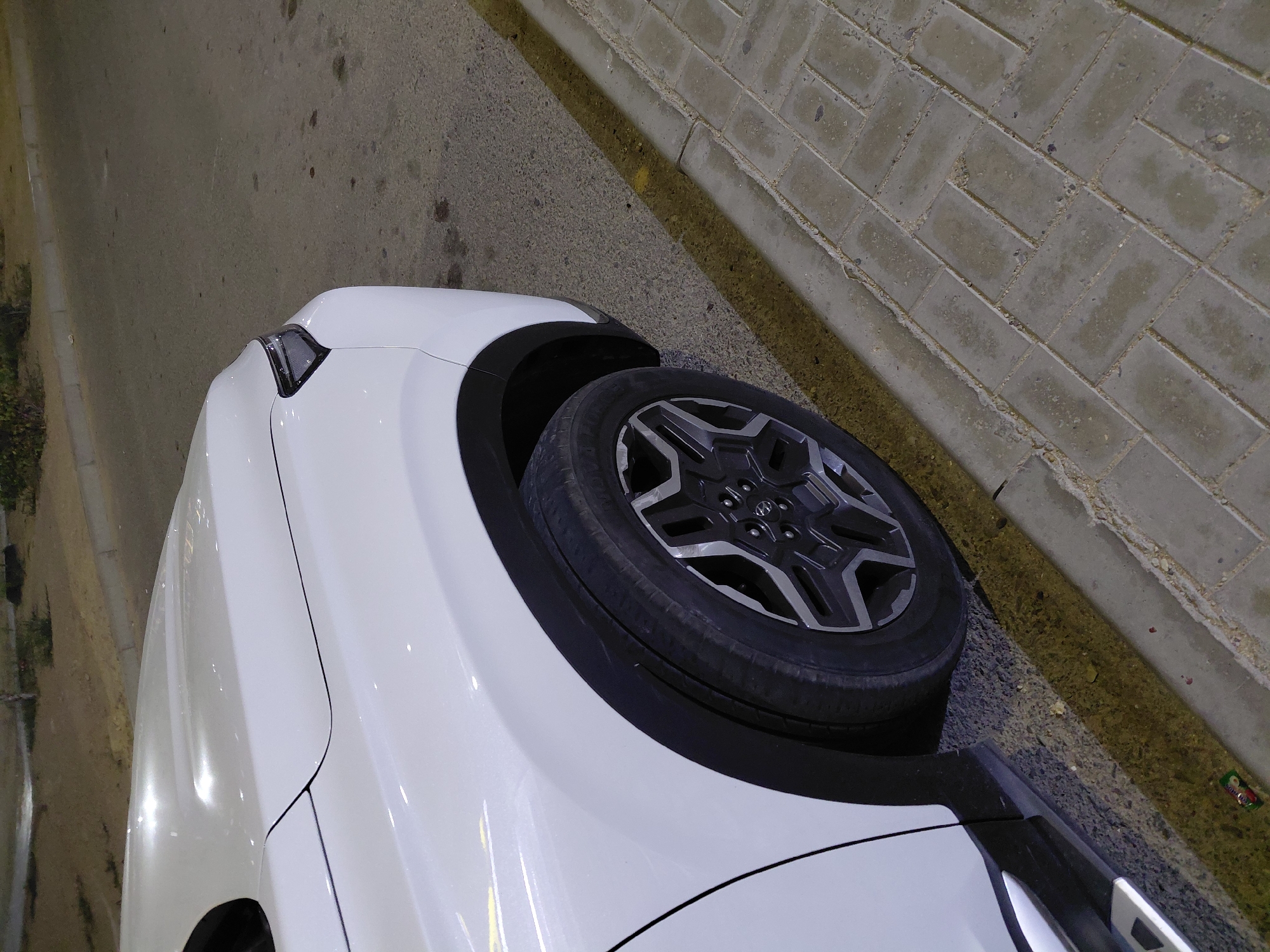 This is a close-up, eye-level, outdoor, medium shot of the front passenger-side wheel and fender of a white SUV. The vehicle is parked on a paved area with a yellow line separating it from a sidewalk made of grey interlocking rectangular pavers. The lighting suggests it is evening or nighttime, with some artificial lights visible in the blurred background, which appears to be a street or parking area with some low vegetation and a concrete curb. The car's tire is black with a silver and black rim. The body of the car is white with a subtle pearlescent sheen. The scene is devoid of people. No specific landmarks or indicators place this image in Aktau, Kazakhstan, beyond the request's context.