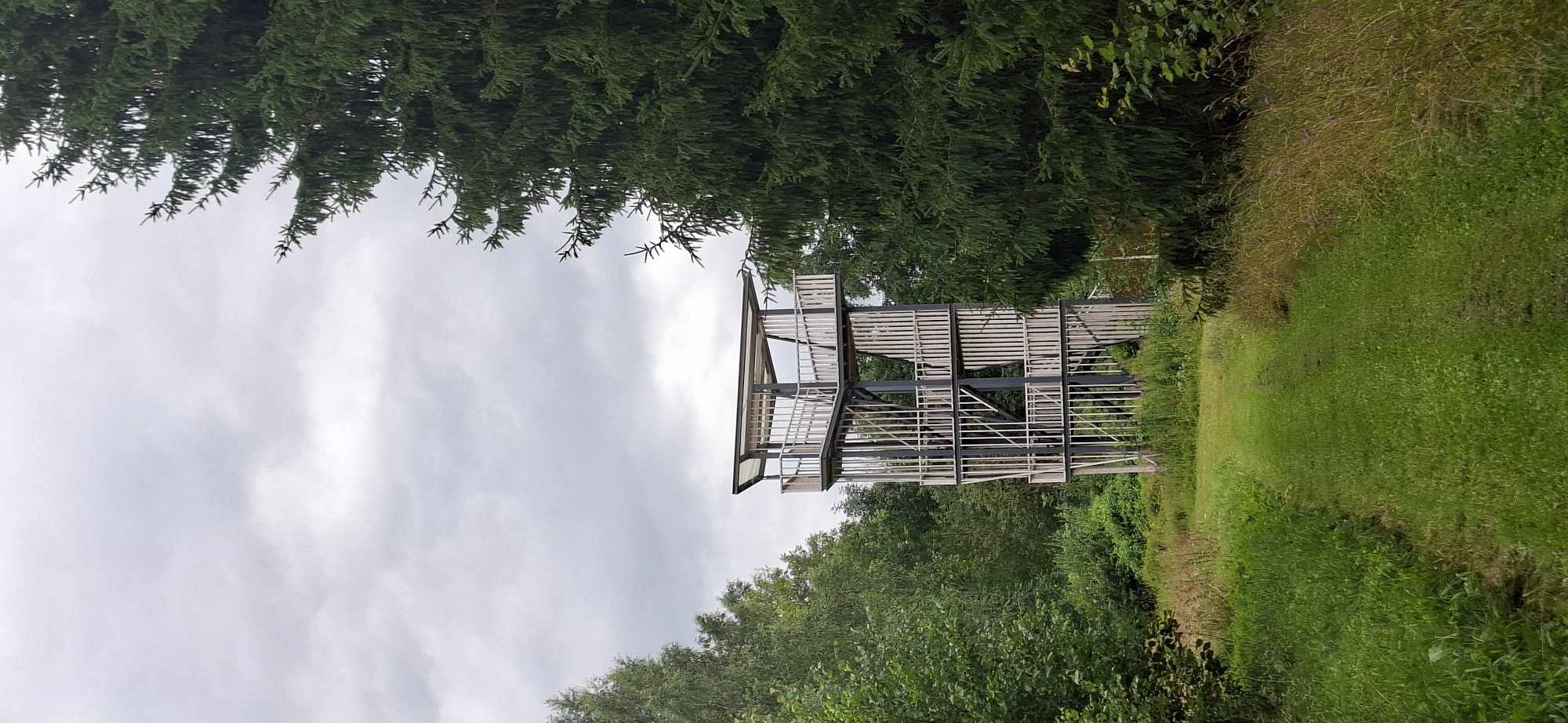 An observation tower, made of metal and wood, stands amidst a forest of tall trees and greenery. The tower is multi-tiered with balconies and a covered top platform. A grassy path leads towards the tower through the vegetation. The sky is overcast with gray clouds. The scene is located in Vihi, Estonia.