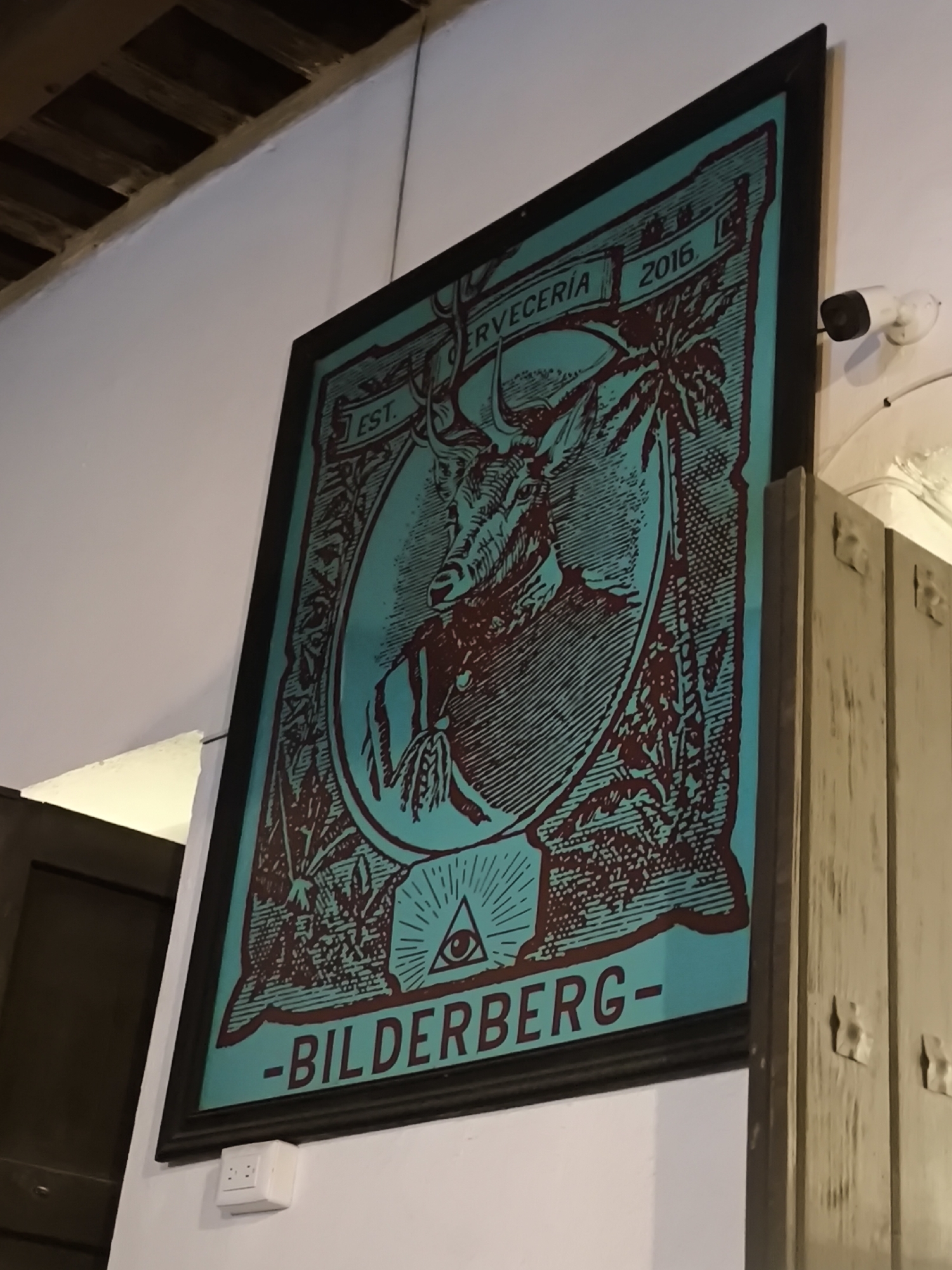 The image shows a framed sign hanging on a white wall. The sign features a teal background with maroon graphics. The graphics depict a deer head inside an oval frame, surrounded by foliage. Above the deer, the sign reads "CERVECERIA" and "2016." Below the deer is an eye-in-pyramid symbol and the word "BILDERBERG." To the left of the sign is a dark brown door. To the right of the sign is a beige-colored wooden structure. A security camera is mounted on the wall above the wooden structure. A white electrical outlet is positioned below the sign. The location is likely Puebla, Mexico, due to the sign's design and the use of the Spanish word "CERVECERIA."