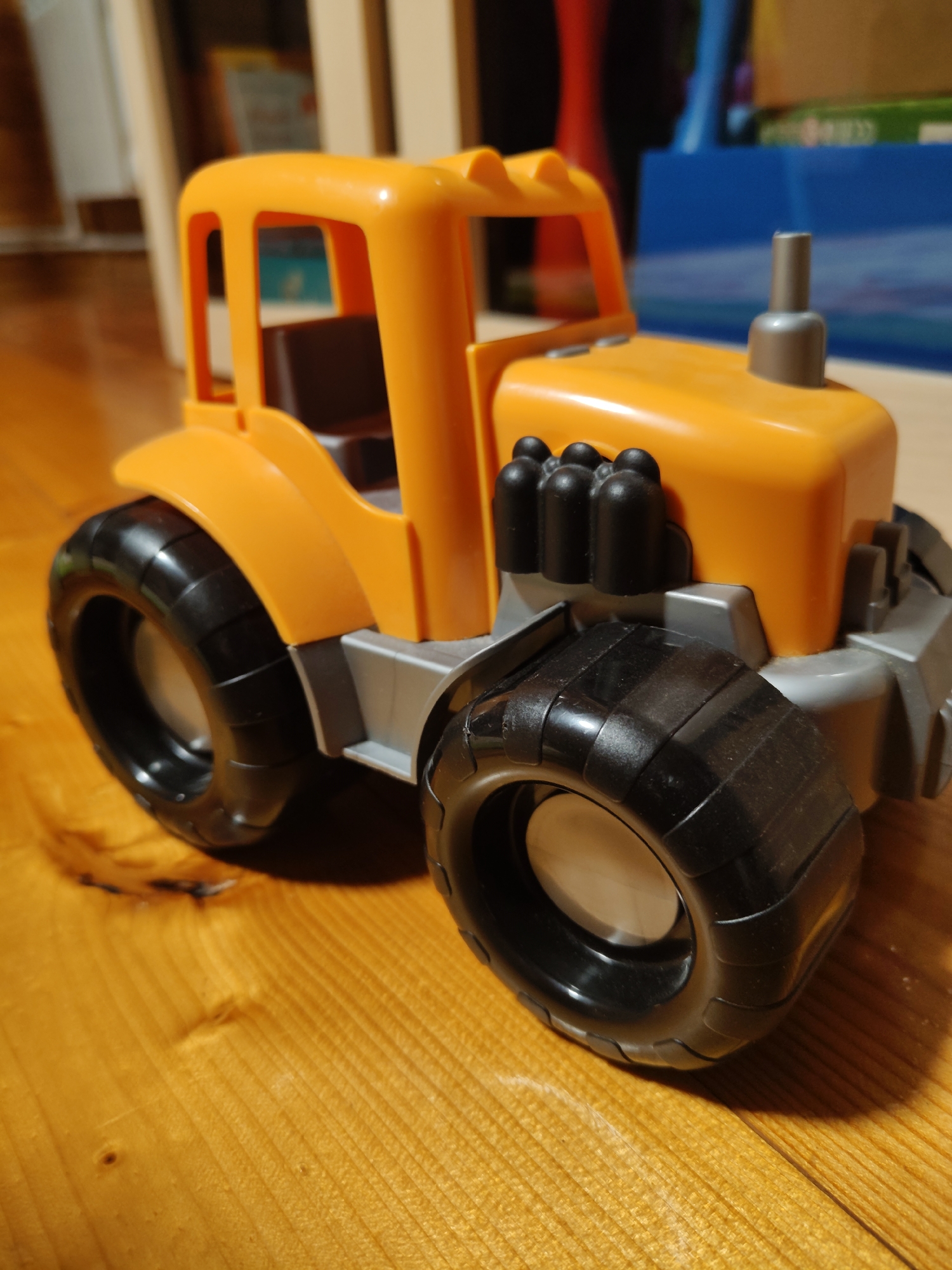 A close-up, eye-level, slightly angled shot shows a bright orange toy tractor with black wheels resting on a polished wooden surface. The tractor is made of plastic and has a grey base. The foreground is dominated by the large, textured rear tire on the right. Behind the tractor, the wooden surface is visible, and further back, a blurry background suggests an indoor setting with some shelving and other objects. The lighting is warm and somewhat dim, creating soft shadows. The scene does not depict any people or specific actions. The location is identified as Фестивальное Снт, Russia, though this information is not visually discernible from the image itself.