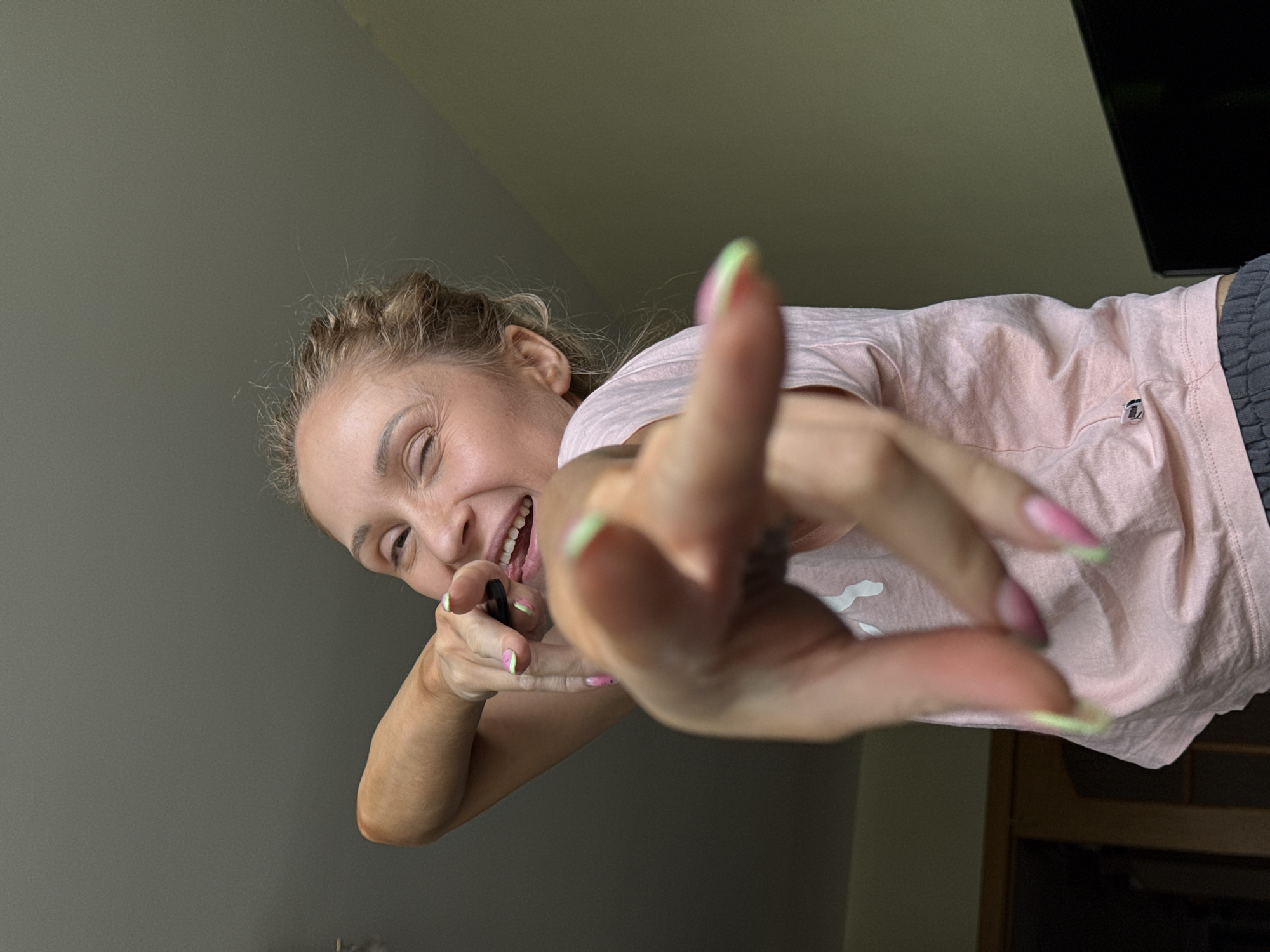 A woman with blonde hair in braids, wearing a pink t-shirt and grey pants, smiles and reaches towards the camera with her right hand, which is in focus. She is holding a black object in her left hand near her mouth. The background consists of gray and light-yellow walls, with a dark object visible in the upper-right corner of the frame. The woman has painted fingernails with a light green edge. The location is not discernible.
