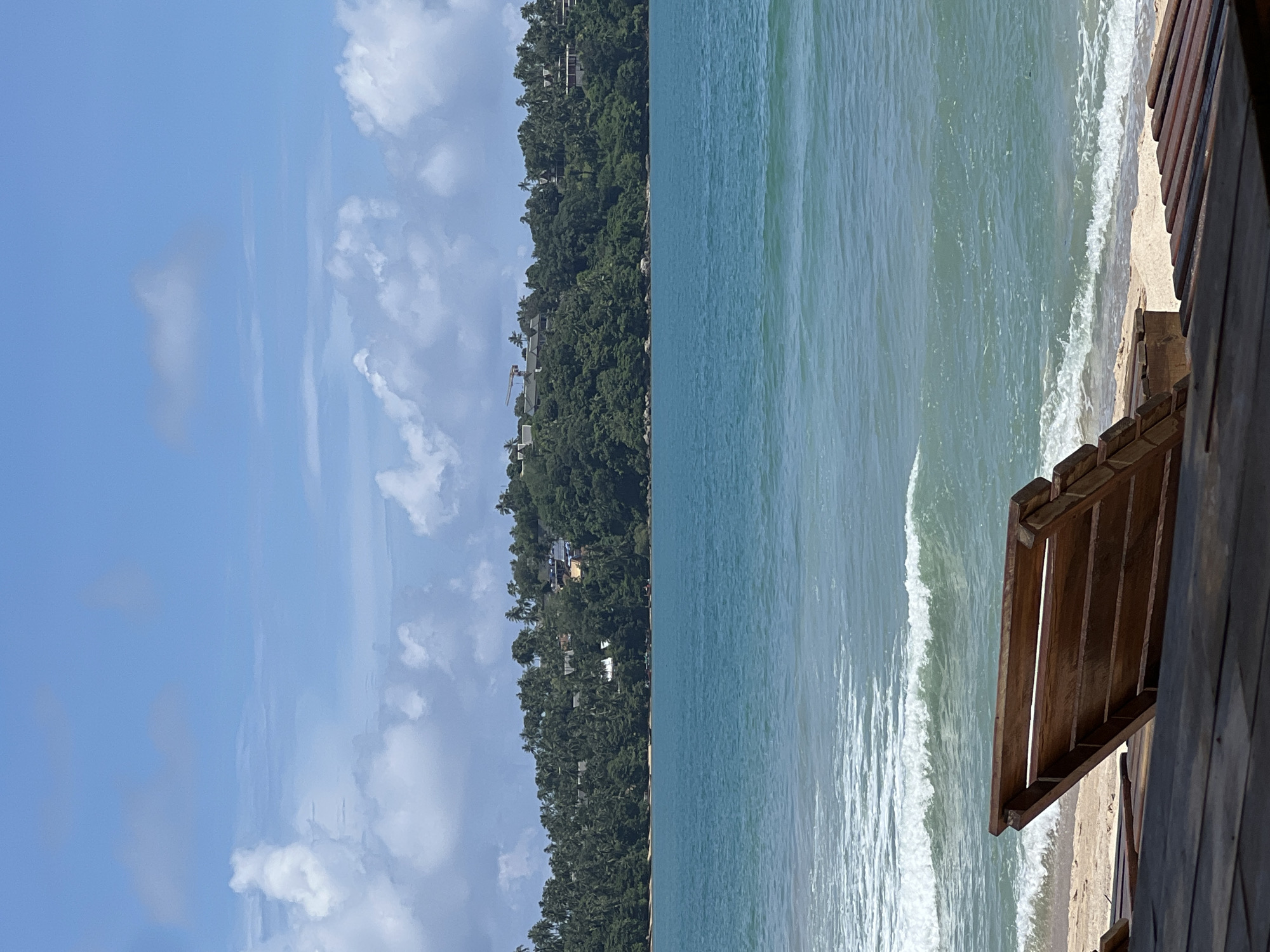A wooden lounge chair sits on a sandy beach, facing a turquoise ocean with gentle waves. In the background, a lush, green, tree-covered hill rises, dotted with buildings and palm trees. The sky above is a clear blue, with scattered white clouds.
