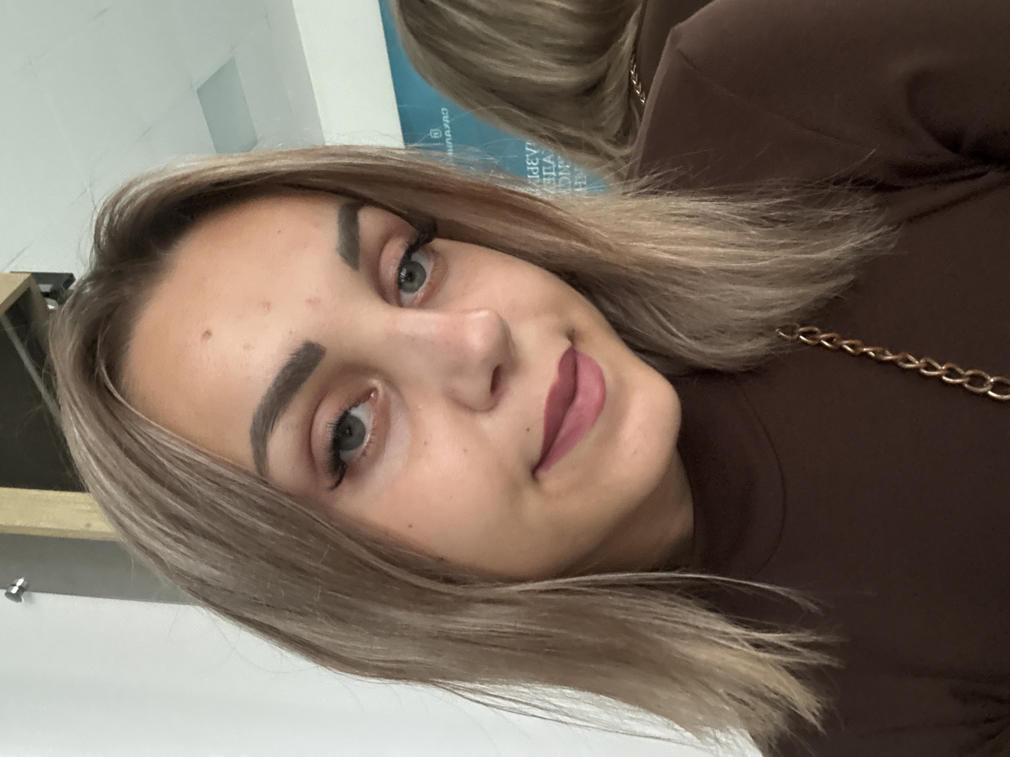 The image is a close-up selfie of a young woman with blonde hair. She is wearing a brown turtleneck shirt with a gold chain detail. Her makeup includes eyeliner and mascara. She is in an indoor environment, possibly an office or public building, given the background which includes a white wall, a wooden beam, and a ceiling light. A reflection of the woman is visible in a mirror behind her, showing the same attire and background elements. The mirror's reflection also shows text on a blue panel, though it is partially obscured and reversed. No specific landmarks or context are provided to identify a precise location within Yuzhno-Sakhalinsk, Russia.