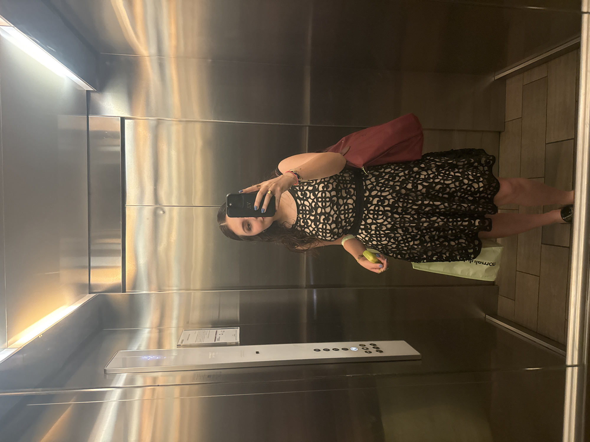 A young adult female is depicted in a full-length mirror selfie within an elevator, located in Mexico City, Mexico. The woman, with long dark hair, holds a dark smartphone with a case displaying white "AV" initials in her right hand, partially obscuring her face, positioned to capture the image. Her right thumbnail is painted blue, and she wears a gold-colored ring. She is dressed in a sleeveless black dress with an intricate, light-colored geometric or floral pattern overlay, and dark flat shoes. She carries a dark red shoulder bag on her left arm and a light green paper shopping bag with white text in her left hand. The elevator interior features brushed stainless steel walls that reflect overhead strip lighting. A white vertical control panel on the left wall contains multiple black floor selection buttons, with one '0' button illuminated, and additional textual information at the top. The elevator floor is composed of rectangular light brown or gray tiles.
