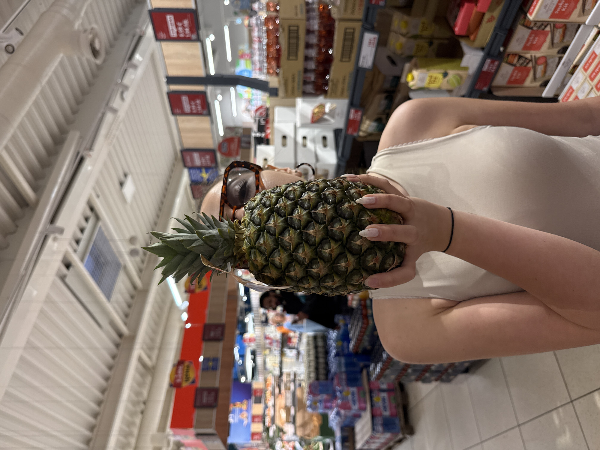 A person holds a pineapple in a supermarket aisle. The person wears sunglasses and a light-colored tank top. The supermarket has shelves stocked with various goods, and signage is visible. The location appears to be a Lidl supermarket in Blagoevgrad, Bulgaria, based on the product displays and store branding.