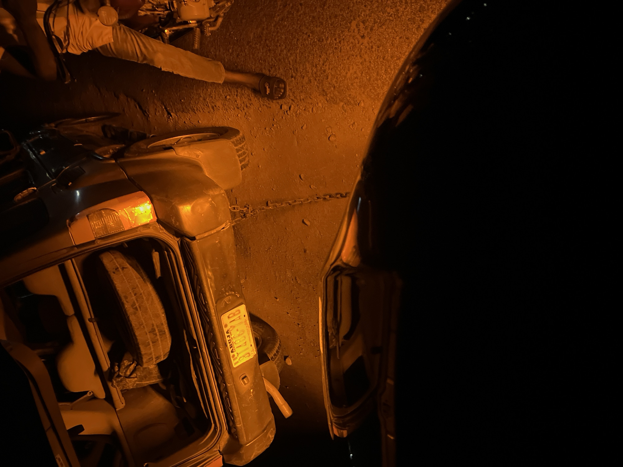 The image depicts a nighttime scene in Kuchingoro, Nigeria. A vehicle, possibly a car, is lying on its side, with the taillight illuminated. Another vehicle is partially visible on the right side of the frame. A man is lying on the ground near the overturned vehicle. A chain is visible on the ground. The license plate of the overturned vehicle reads "ABUJA", and the plate number is partially visible as "118G...". The environment appears to be a street or roadside.