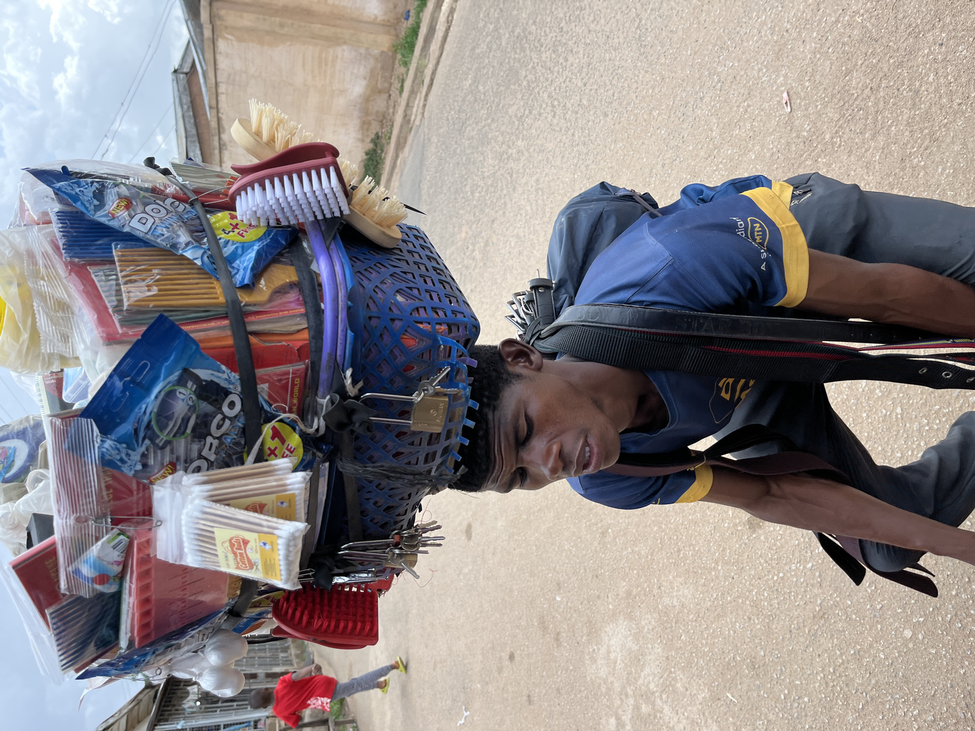 A street vendor in Jos, Nigeria, carries his wares stacked high on his head in a blue plastic basket, secured with black straps, walking on a paved road. He is a young, dark-skinned man wearing a blue shirt with yellow trim and dark pants. The basket contains various household items, including brushes, cleaning supplies, cotton swabs, and small plastic containers. Padlocks and keys are attached to the basket's rim. A backpack sits on his back, underneath the basket. The background shows buildings and greenery along the roadside, with a cloudy sky above. A person wearing a red shirt and grey pants walks in the lower-left corner of the image.