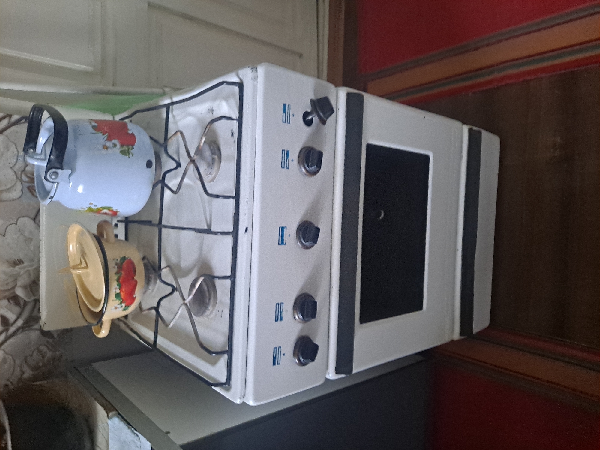 A white gas stove is positioned in a room. On top of the stove, there is a light blue kettle with red floral patterns and a yellow pot with a lid adorned with red fruit images. The stove has four burners, and the control knobs are visible on the front panel. The floor beneath the stove is covered by a rug with horizontal stripes in red, orange, and beige. The background includes a section of a white door frame and patterned wallpaper. The scene appears to be an interior setting within a home, possibly a kitchen.