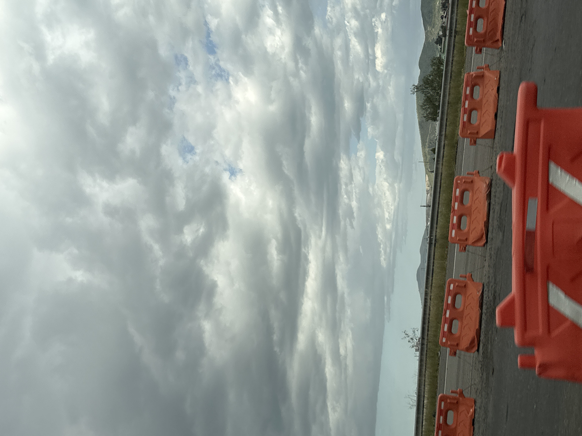This is a vertical, medium shot photograph of a road in Santa Clara Ocoyucan, Mexico, under a cloudy sky. In the foreground, several bright orange plastic water-filled barriers, some with reflective white stripes, are positioned on the asphalt. In the middle ground, a line of identical orange barriers stretches across the road, indicating a construction zone or road closure. Beyond the barriers, a grey metal guardrail runs parallel to the road, with sparse green vegetation and distant, rolling hills under a heavy, overcast sky. The clouds are thick and grey, with patches of lighter grey and white. The overall lighting is dim and diffused due to the cloud cover.