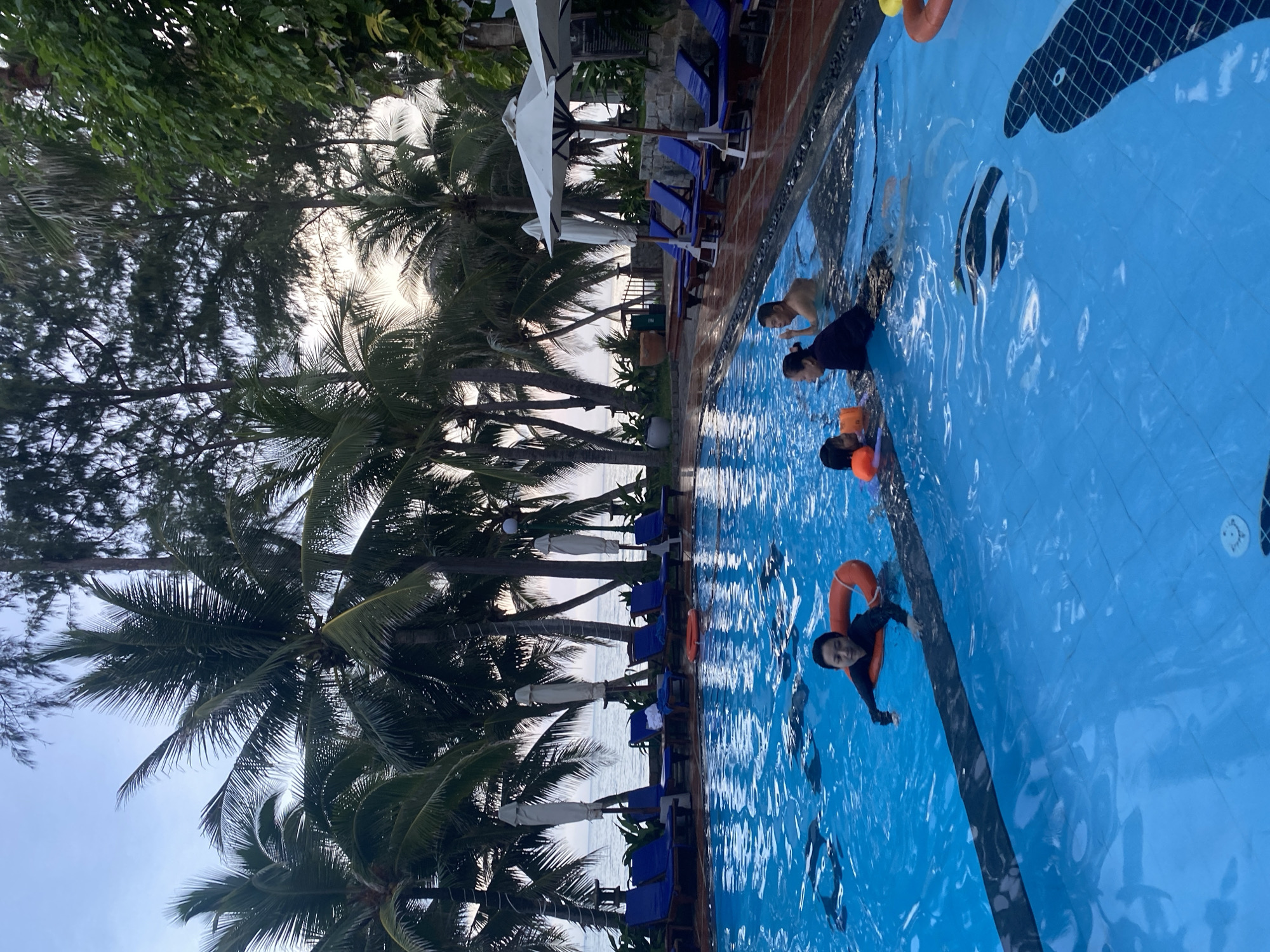 A group of people are enjoying a swimming pool in Phu Thuy Ward, Vietnam. The pool is outdoors and surrounded by palm trees, with lounge chairs and umbrellas visible in the background. Several people are in the water, some wearing life preservers. The sky is overcast, suggesting it may be early morning or late afternoon. The scene appears to be a vacation resort or a private residence.