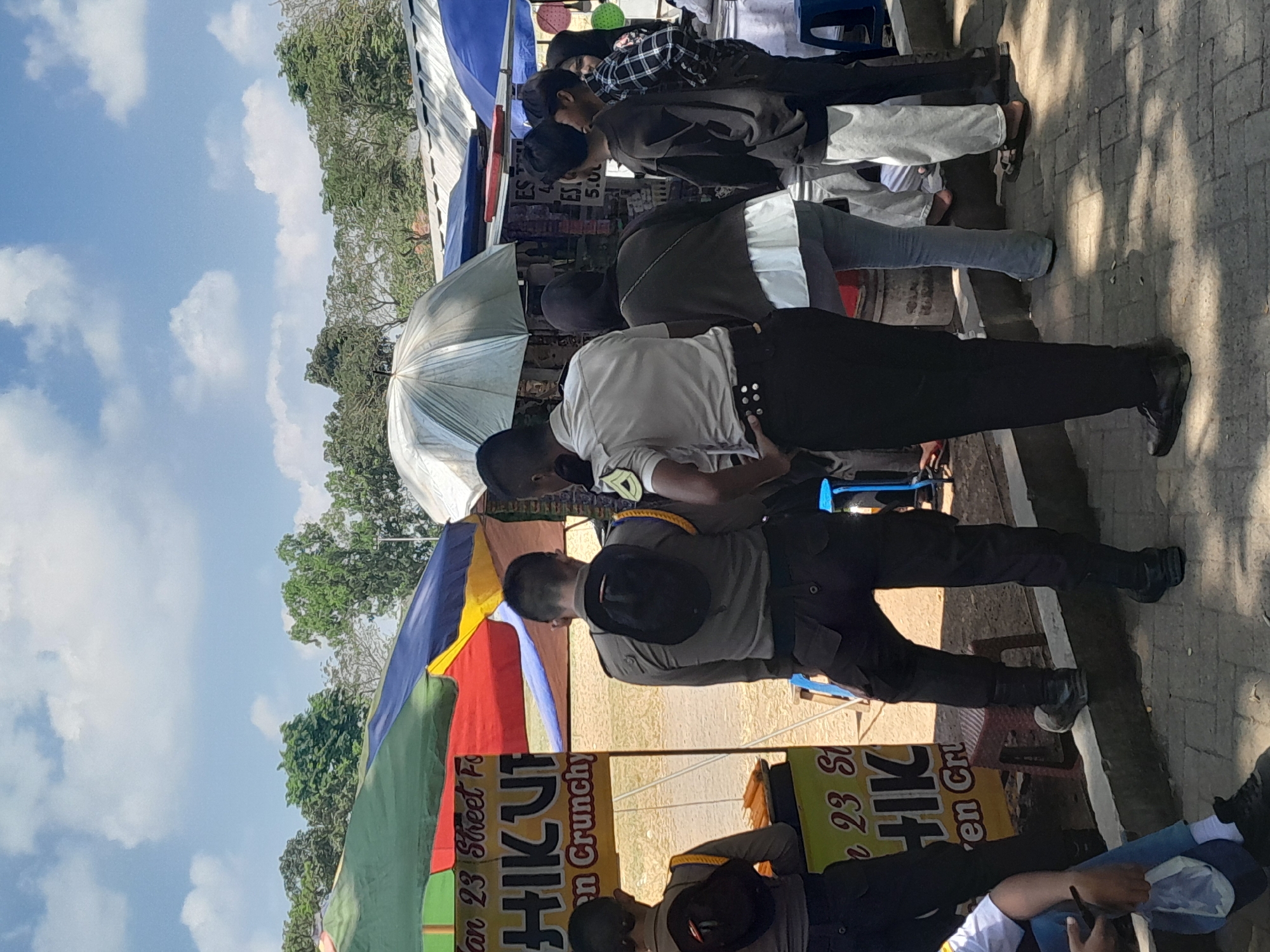 In Genteng Kulon, Indonesia, under a bright, cloudy sky, a group of people are gathered near market stalls. Two individuals in what appear to be law enforcement uniforms, distinguished by their dark pants and shirts with yellow epaulets, are standing near a food stall. One of the uniformed individuals is facing away from the camera, his hat perched on his hip, while the other is turned slightly, observing the surroundings.
The food stall features a colorful awning with red, blue, green, and yellow sections. A sign on the stall reads "an 23 Street Fo" and "HIK UP en Crunchy". In front of the stall, several skewers of food, possibly corn dogs, are being displayed.
Further down the row of stalls, an open, silver umbrella provides shade over a display. Several other individuals are seen browsing or interacting in this area. One person, wearing a dark long-sleeved shirt, is looking towards the stalls. Another person in a light-colored top and gray pants is also present, as is someone in a black hoodie and another in a plaid shirt. The ground is paved with bricks, and a concrete curb separates the paved area from a dirt or sandy patch where the stalls are set up. The overall atmosphere appears to be that of a local outdoor market or street fair.
