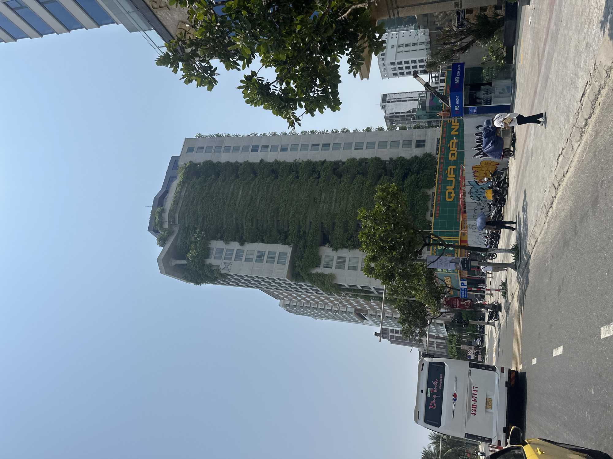 The image depicts a street scene in Đà Nẵng, Vietnam. A tall building covered in green vines is prominently featured. A white minibus with license plate "43H1-7147" is visible on the road. Signage indicates a parking area. People are seen walking on the sidewalk next to parked motorbikes. The signage includes references to "QUÀ ĐÀ NẴNG," a local shopping center, and ATMs.