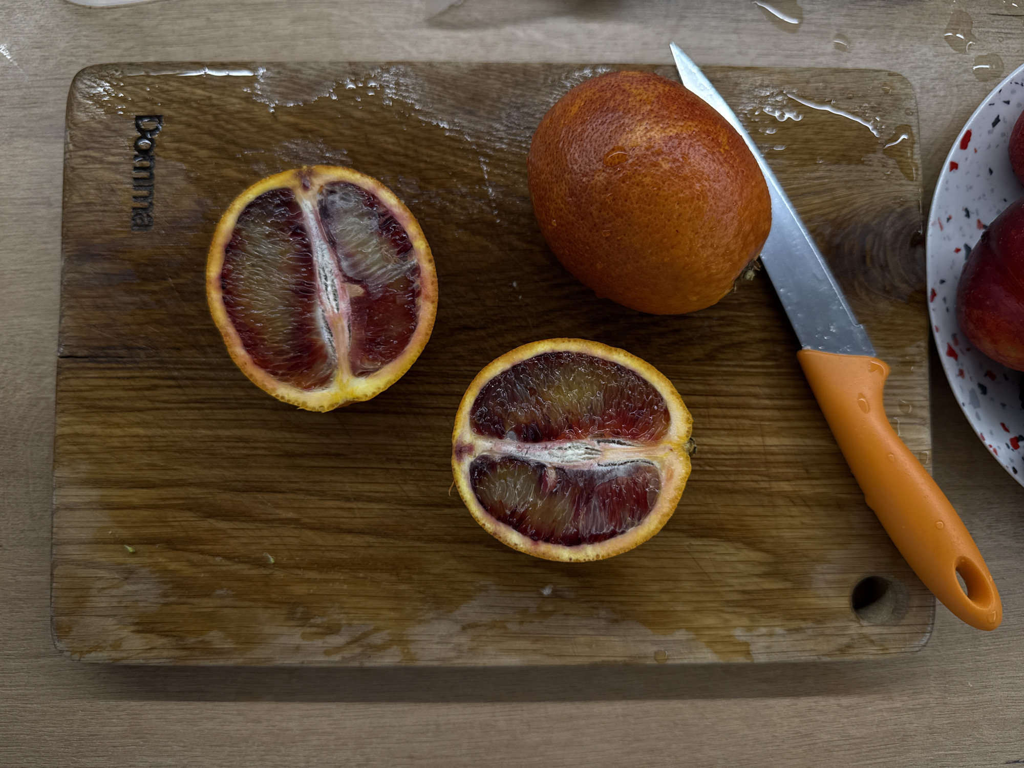An overhead shot captures a wooden cutting board on a light wooden or laminate countertop, featuring blood oranges and a knife. Two halves of a blood orange are positioned on the left side of the cutting board, revealing their vibrant red and orange interior. A whole blood orange rests to their right, beside a silver knife with a distinct orange handle. The cutting board exhibits various splashes and droplets of water or juice, indicating recent activity.
The word "Bonna" is vertically visible, engraved or printed, in the upper left corner of the cutting board. A circular hole is present in the bottom right corner of the cutting board, near the knife's handle. On the far right, part of a white bowl with red and black speckles is visible, holding what appears to be additional fruits. The scene depicts an indoor setting, likely a kitchen, centered around the preparation of fruit. No individuals are present in the image.
