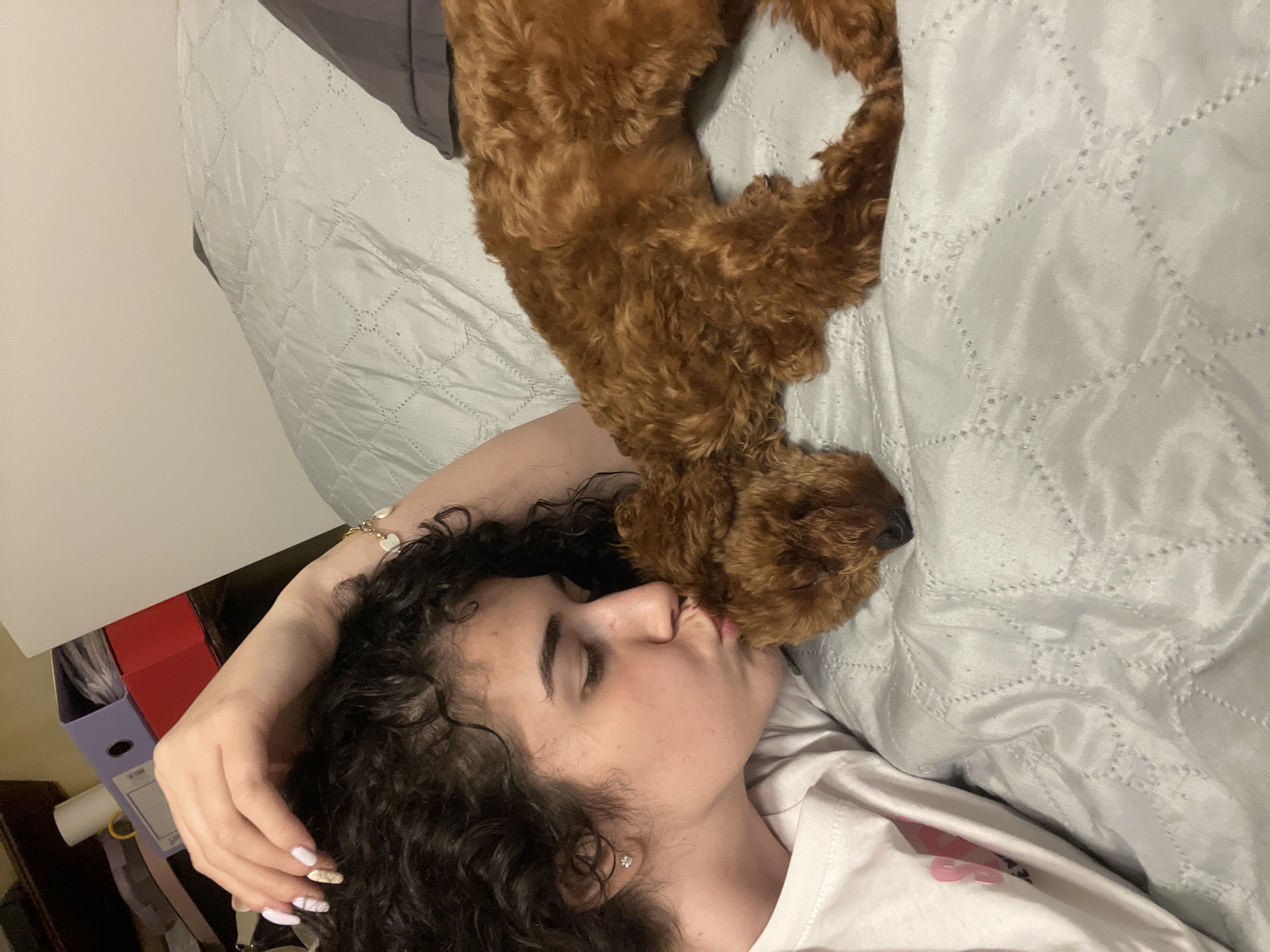 A young woman lies on a grey quilted bed cover, eyes closed and facing left. A reddish-brown dog rests its head near the woman's face. The woman wears a white t-shirt and has curly dark hair. Her left arm is raised near her head. To the left of the bed is a desk or shelf with stacked items including a red box and a purple binder. It is not possible to identify the location in Sofia, Bulgaria based on the image.