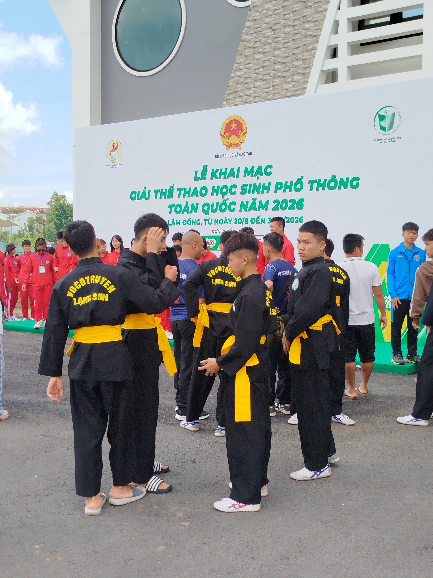 A group of young people, some in martial arts uniforms, stand in front of a large white banner. The banner reads "LỄ KHAI MẠC GIẢI THỂ THAO HỌC SINH PHỔ THÔNG TOÀN QUỐC NĂM 2026 LÂM ĐỒNG, TỪ NGÀY 20/6 ĐẾN 30/2026". This indicates the opening ceremony of the National High School Sports Competition 2026 in Lam Dong province, taking place from June 20th to June 30th, 2026. The building in the background appears to be a modern structure with a large circular window. The ground is paved and there are some trees visible in the distance, suggesting an outdoor event. The individuals in the foreground, wearing black uniforms with yellow sashes, are likely participants or instructors in a martial arts discipline. Some are engaged in conversation or practice, while others look towards the camera. The overall atmosphere seems to be festive and organized.