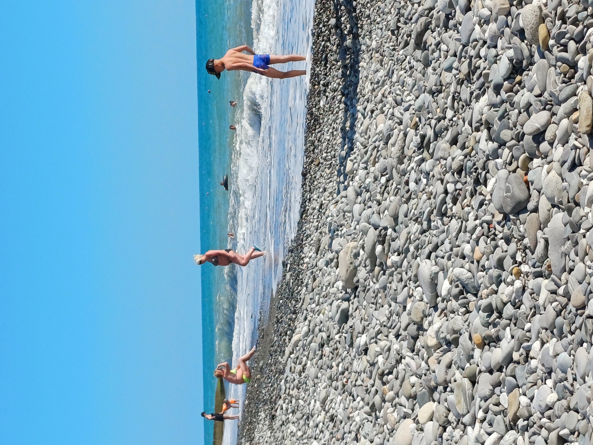 The image depicts a sunny day at a pebble beach in Sochi, Russia. The foreground is filled with smooth, grey pebbles of various sizes. In the midground, several people are visible. A young boy wearing blue swim trunks and a black baseball cap stands at the edge of the water, facing away from the camera. Further down the beach, closer to the waterline, a woman with blonde hair in a black bikini walks towards the sea. To her left, a woman in a yellow bikini is sitting on the wet pebbles, facing the sea. In the distance, the turquoise sea meets a clear blue sky, with small waves breaking on the shore. A few other people are visible in the water, some appearing to be swimming. The atmosphere is one of leisure and recreation on a warm summer day.