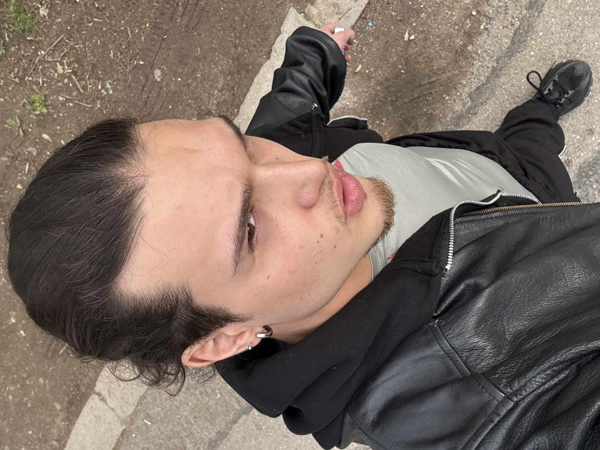 An overhead, tilted portrait features a person outdoors, looking up and to the right with a neutral expression. The subject, appearing to be a young male, has dark hair styled back, showcasing a visible hairline and some hair regrowth. Light stubble or a short beard covers the chin and mustache area. A faint reddish mark is present on the forehead. The left ear displays two earrings: a silver hoop and a smaller stud or ring.
The person is dressed in a black leather jacket worn over a black hooded sweatshirt. A light-colored t-shirt is visible beneath the layers. Black pants and black athletic shoes complete the outfit. In the upper right corner of the image, one hand is partially visible, holding a small, light-colored object, possibly a vape pen or cigarette.
The background consists of a textured ground surface, a mix of dirt, gravel, and sections of concrete or asphalt. Sparse green weeds, small twigs, and white specks of debris are scattered across the ground. A concrete curb or a painted white line runs diagonally through the lower part of the frame. The lighting suggests it is daytime, with soft, diffused light. There is no visible text in the image.