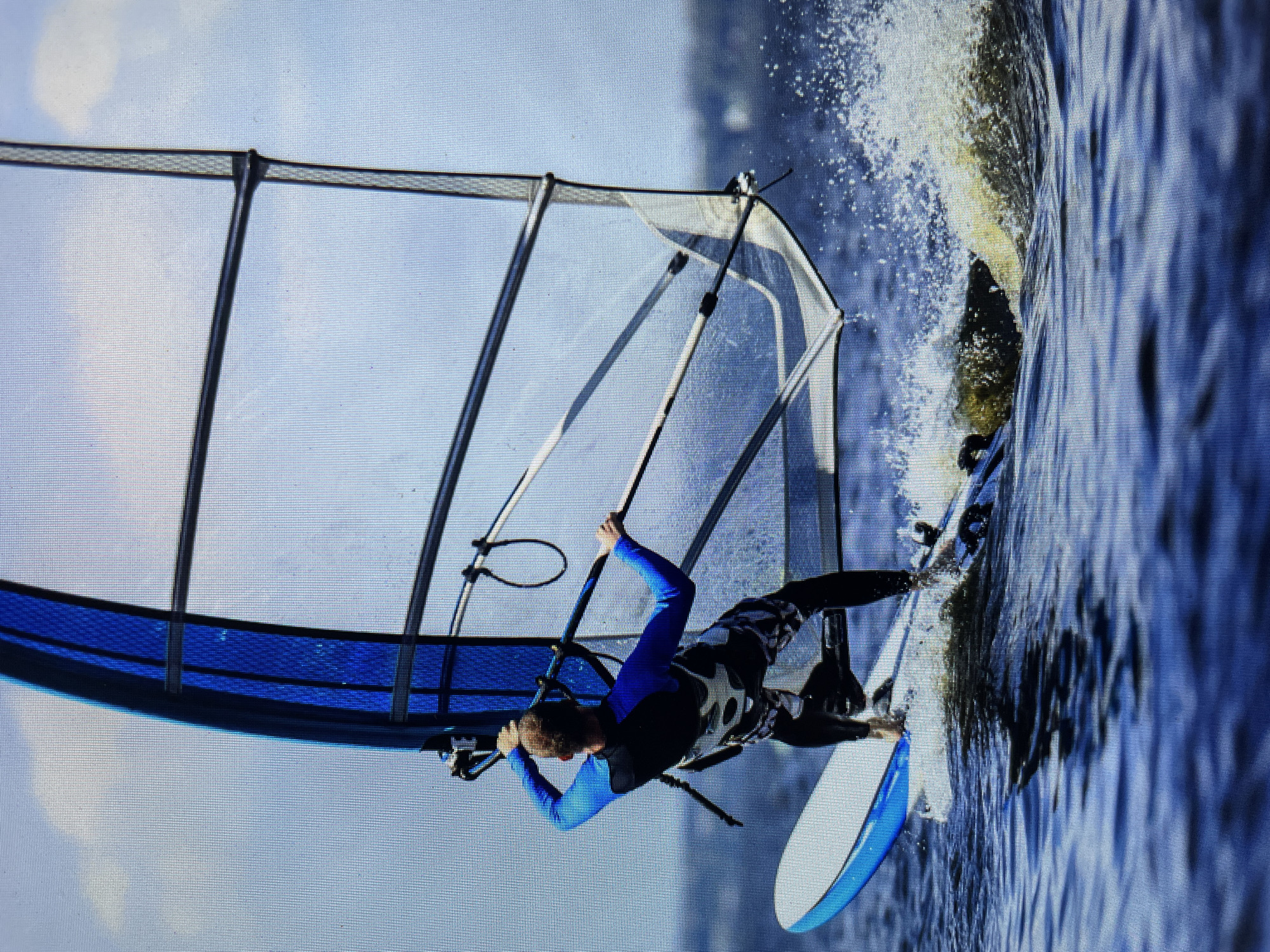 A man windsurfing in the waters off Bacong, Philippines. The man, wearing a wetsuit, is standing on the windsurfing board, holding onto the sail. Splashes of water are visible where the board cuts through the water. The environment is an expanse of water under a bright sky.