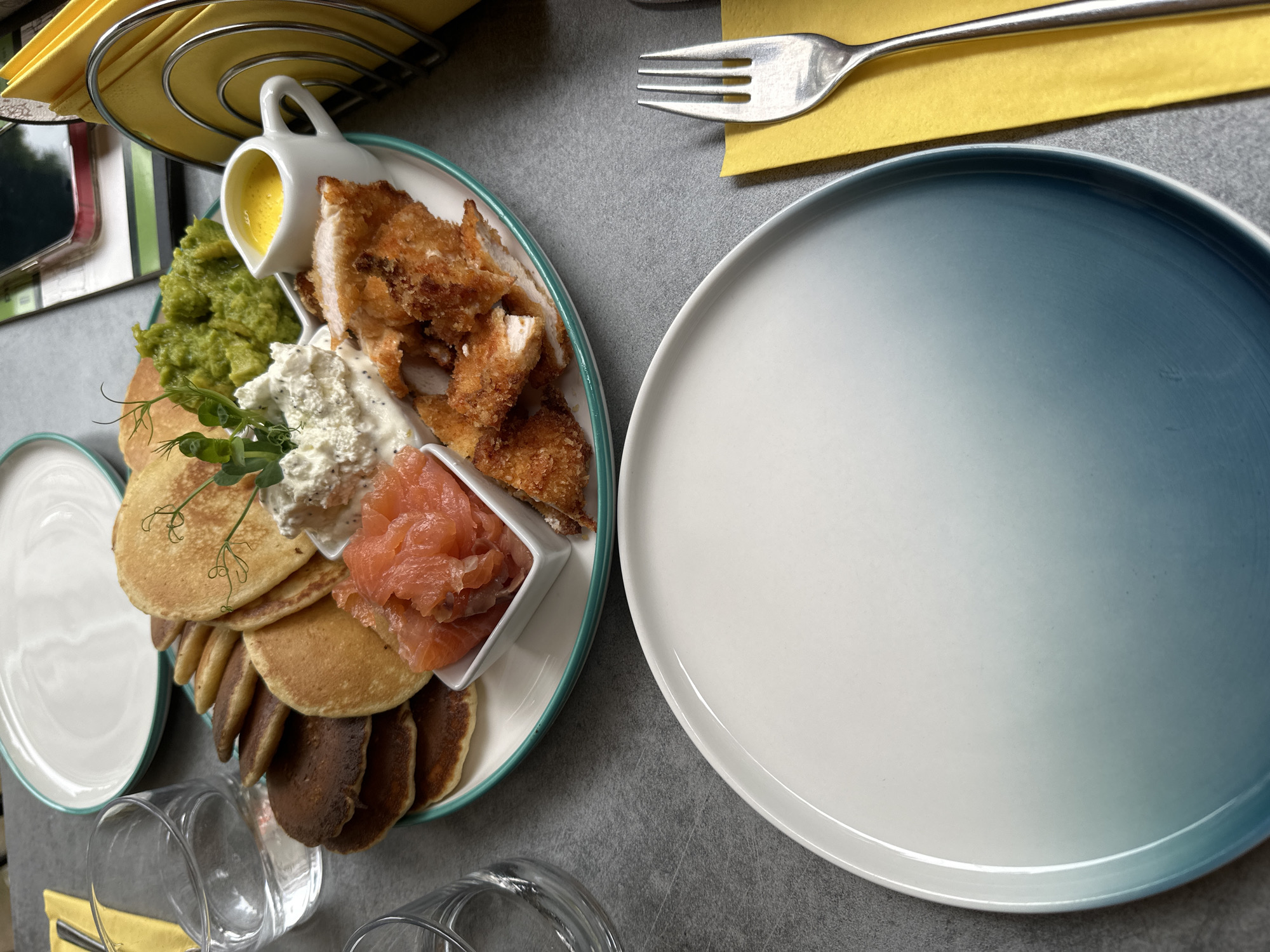 In Sofia, Bulgaria, on a gray textured table, a brunch dish is presented on a white plate with a teal rim. The dish features a stack of small, golden-brown pancakes, accompanied by a small white bowl filled with pink smoked salmon. Next to the salmon is a dollop of white cream cheese with black flecks, topped with delicate green sprouts. Adjacent to this is a mound of vibrant green mashed peas. A small white pitcher holds a bright yellow sauce, possibly hollandaise. To the right of these elements are several pieces of golden-brown, breaded chicken tenders. In the foreground, a large, empty white plate with a gradient blue rim sits ready for food. A glass of water is visible on the left, and a yellow napkin with a fork is on the right. A napkin holder with yellow napkins is in the background, along with a smartphone.