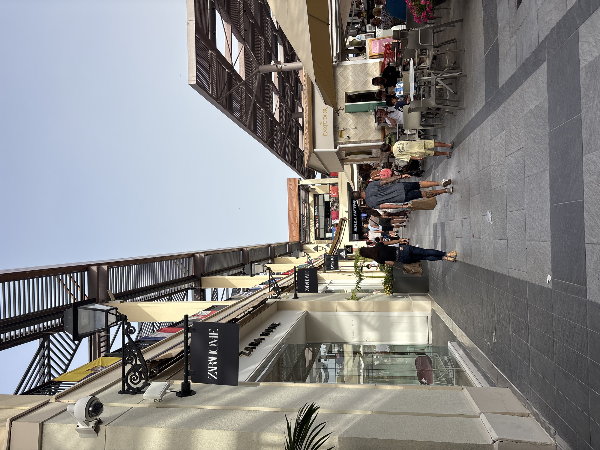 This is an eye-level, vertical, full shot of a pedestrian walkway in a retail area, likely an outdoor shopping center, under a clear sky. The architecture features light-colored building facades on the left and a structure with a slatted roof overhang on the right, creating a covered walkway. Various shops are visible, including "ZARA HOME" with its signage prominently displayed, as well as other stores identified by signs for "OYSHO" and "SKECHERS". In the foreground, several people are walking on a paved walkway made of large rectangular tiles. On the left, a woman in dark jeans and a black shirt walks away from the camera. In the center, a man in a gray t-shirt and dark shorts walks away from the camera, followed by a young boy in a light yellow t-shirt and shorts. Further down the walkway, groups of people are seated at tables outside a cafe identified by a "CARTE D'OR" sign. The overall impression is of a bustling commercial area on a sunny day, possibly in Orihuela, Spain, given the context provided.