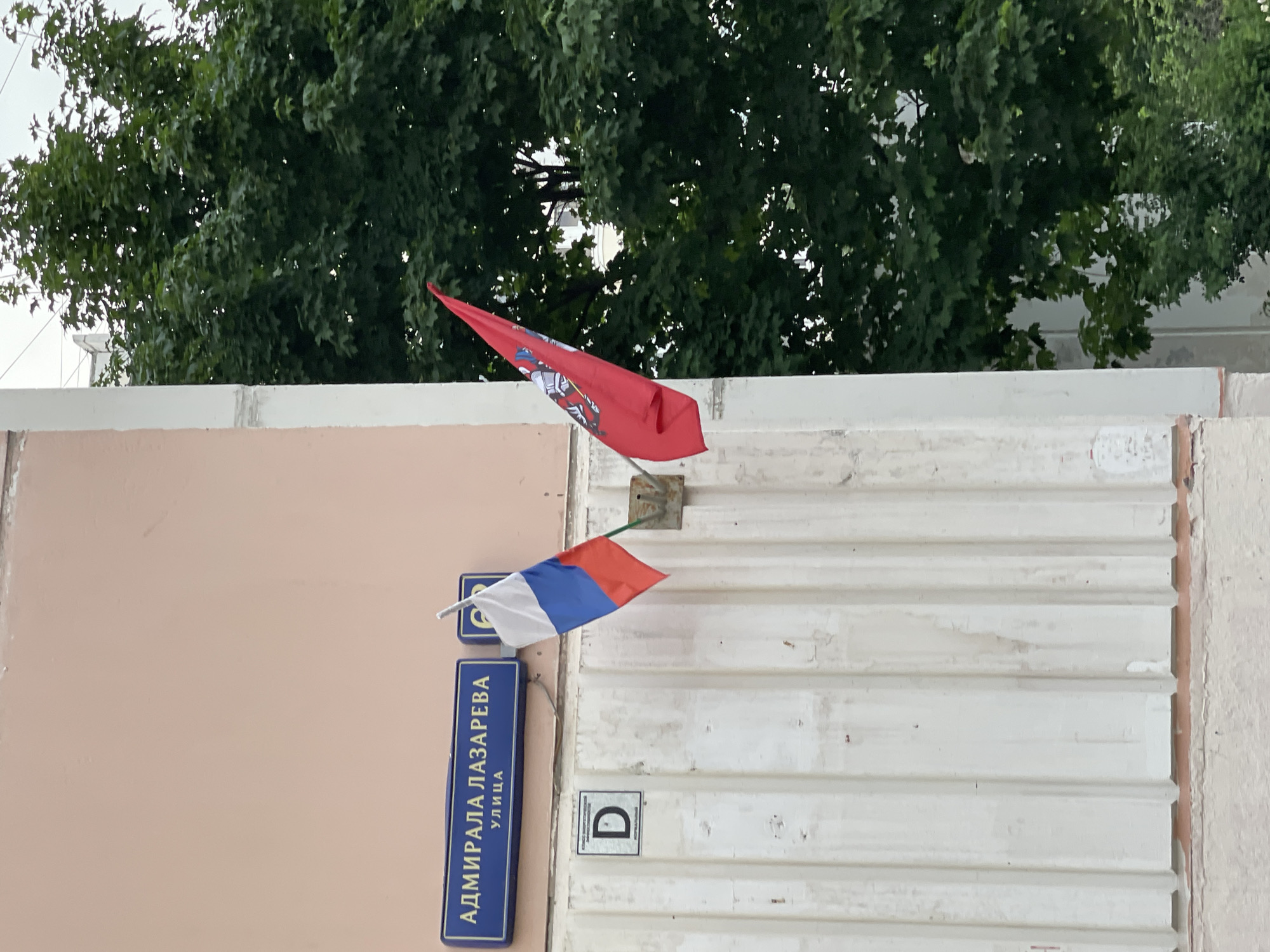 The image shows a close-up of a building exterior with two flags displayed. On the left, a Russian flag, consisting of three horizontal stripes of white, blue, and red, is attached to a metal pole. To the right of the Russian flag, a red flag with a coat of arms is also displayed. Above the flags, a blue street sign reads "АДМИРАЛА ЛАЗАРЕВА УЛИЦА," indicating Admiral Lazarev Street. Below the street sign, a white square plaque with the letter "D" in black is visible. The building has a pink facade on the upper part and a white, ribbed lower section. A lush green tree fills the right side of the frame, partially obscuring buildings in the background. The overall scene appears to be an outdoor urban environment.