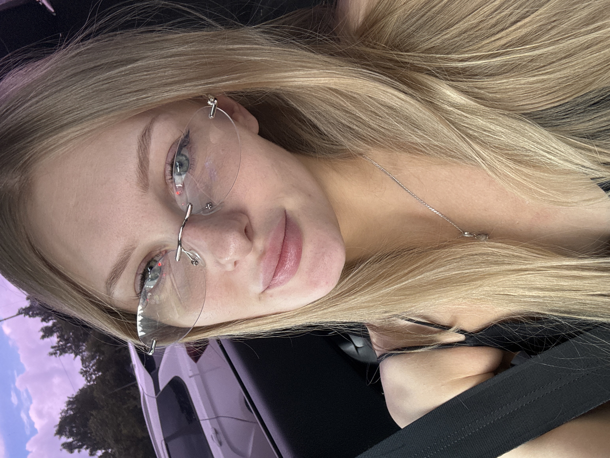 A close-up, horizontally oriented selfie shows a young woman with blonde hair and glasses. She is wearing a black top and a seatbelt. Her eyes are blue, and she has a light complexion with freckles. She is wearing rimless glasses, and a thin silver necklace is visible. The background appears to be inside a vehicle, with a hint of the sky and trees visible through the window. The sky has purple and pink hues, suggesting it might be dusk or dawn. The woman's expression is relaxed and pleasant. There is no visible text in the image. The location context is Perm, Russia.