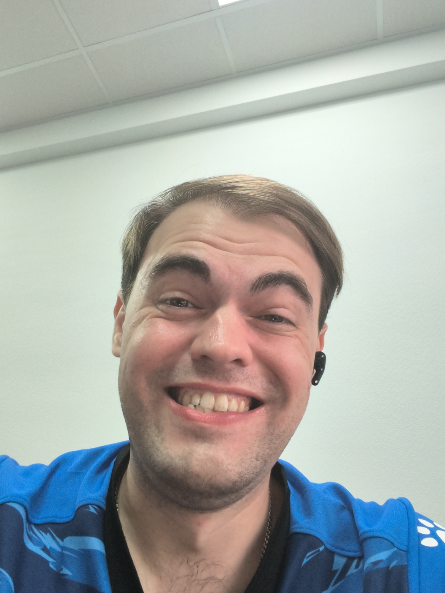A close-up, waist-up selfie shows a Caucasian man with light brown hair and fair skin, smiling broadly and looking directly at the camera. He is wearing a bright blue shirt with a dark blue and white pattern. A small black earbud is visible in his right ear. The background is a plain, light-colored wall with a white ceiling panel overhead. The lighting is bright and appears to be from overhead sources. The man's face is slightly flushed, and he has a few light wrinkles around his eyes and forehead, indicating a happy expression. The overall impression is of a casual and friendly demeanor. The location is not specified in the image itself, and therefore cannot be determined.