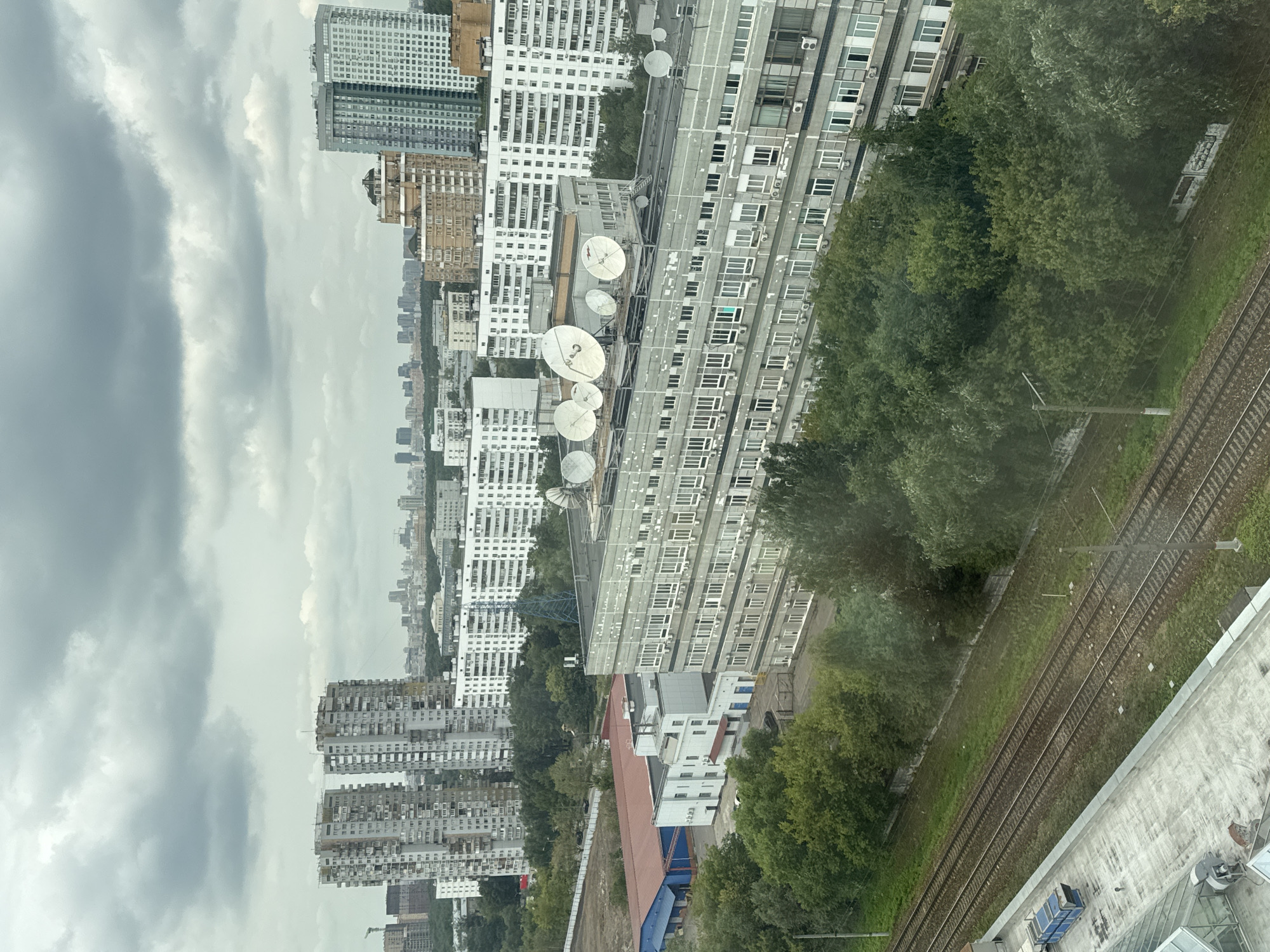 The image shows an aerial view of a cityscape with overcast skies. In the foreground, there are train tracks running diagonally across the frame, bordered by lush green trees on one side and a concrete structure with some industrial equipment on the other. Further into the city, there is a large, grey, multi-story building with numerous windows and air conditioning units. On top of this building, several satellite dishes are visible. Beyond this, a dense collection of residential buildings of varying heights and architectural styles dominate the mid-ground and background. Some of these buildings are modern high-rises, while others appear to be older, Brutalist-style apartment blocks. The general impression is of a densely populated urban environment with a mix of residential and industrial structures. The presence of these architectural styles, particularly the Brutalist blocks and the overall layout, is characteristic of some areas in Khimki, Russia, a city known for its large Soviet-era residential districts and proximity to Moscow.