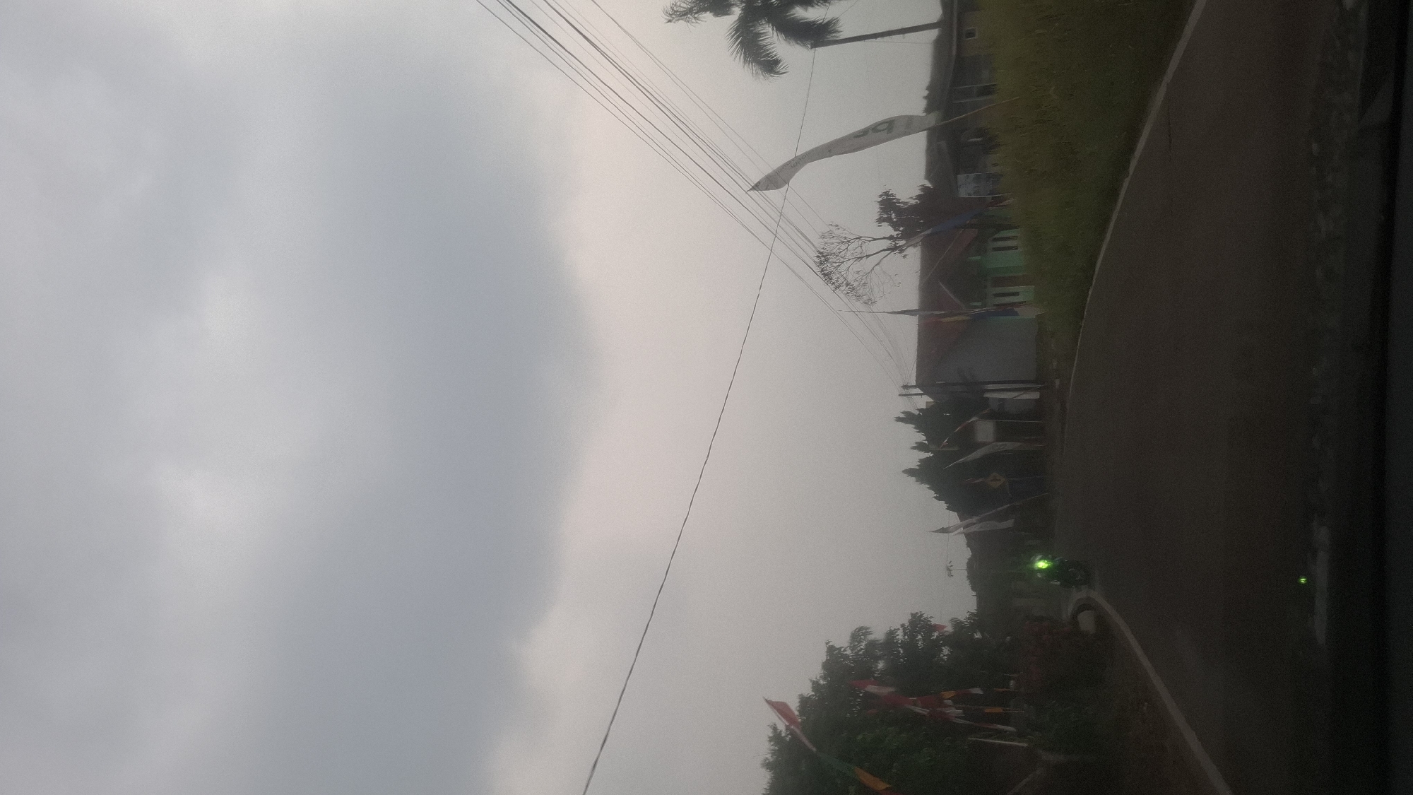 The image captures a slightly overcast day in what appears to be a rural or suburban area. A paved road curves through the scene, with a single motorcycle visible in the distance, its headlights on. Along the roadside, several houses with red-tiled roofs and simple facades are present, some adorned with flags. Green foliage, including trees and grass, lines the road. Electrical wires are strung across the sky, supported by poles. The overall atmosphere suggests a quiet, everyday scene in Cibinong Hilir, Indonesia.