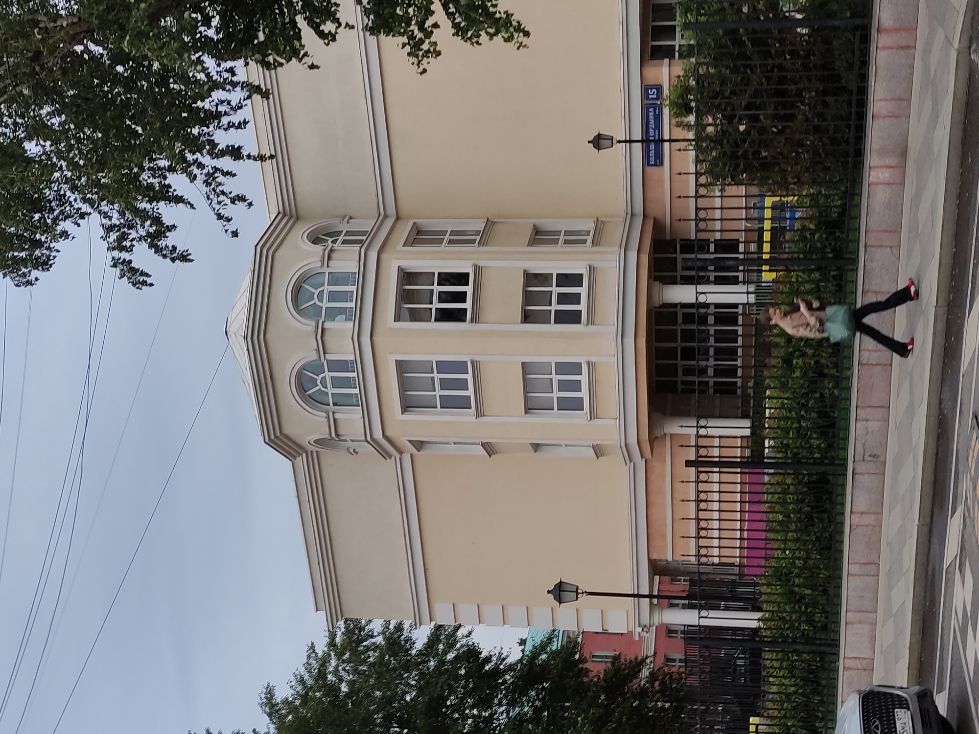 The image depicts a building in an urban environment. The building is painted in a light yellow color with white window frames and trim. It features a distinctive architectural style with arched windows on the top floor. A blue street sign with white lettering, reading "Большой Ордынка 15", is visible on the side of the building, indicating the street name and number.
A black metal fence runs along the sidewalk in front of the building, partially obscuring a person who is walking. The individual is wearing dark pants and red shoes. The sidewalk is paved with a checkered pattern of light and dark tiles.
Trees with green foliage are present on both sides of the building, partially framing the scene. Overhead, utility lines are visible against the sky. The environment suggests a cloudy day.