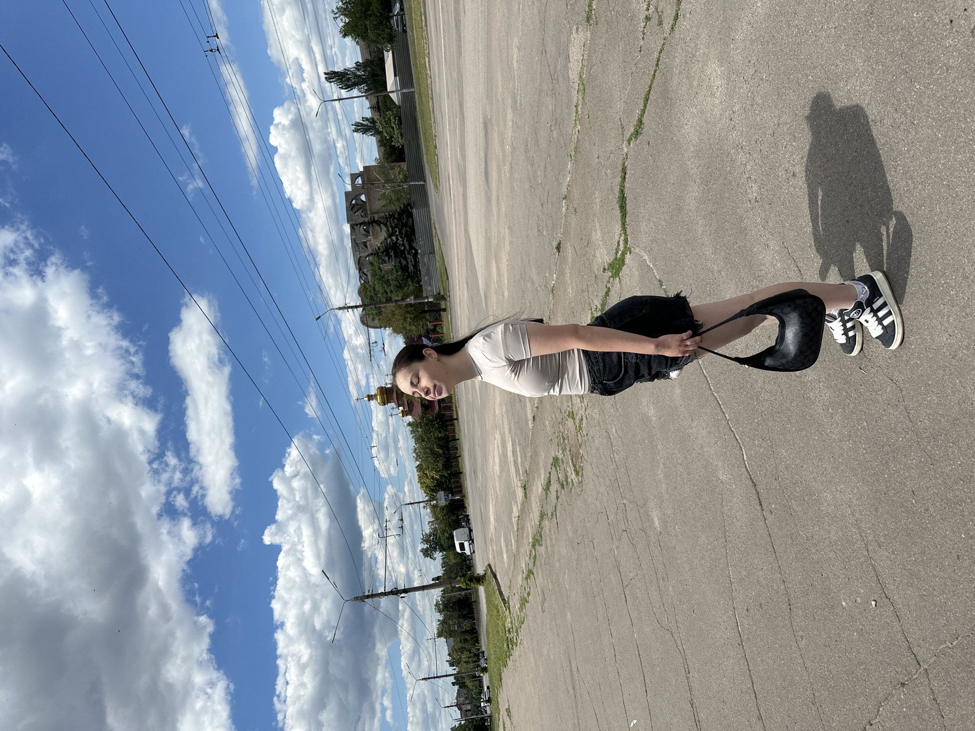 A young woman stands on a cracked asphalt surface, looking up at the sky. She is wearing a light-colored t-shirt, a black denim skirt, and black and white athletic shoes. A black handbag hangs from her right hand. In the background, there are power lines, streetlights, trees, buildings, and vehicles on a road. A golden-domed building, likely a church, is visible in the distance. The sky is blue with scattered white clouds.